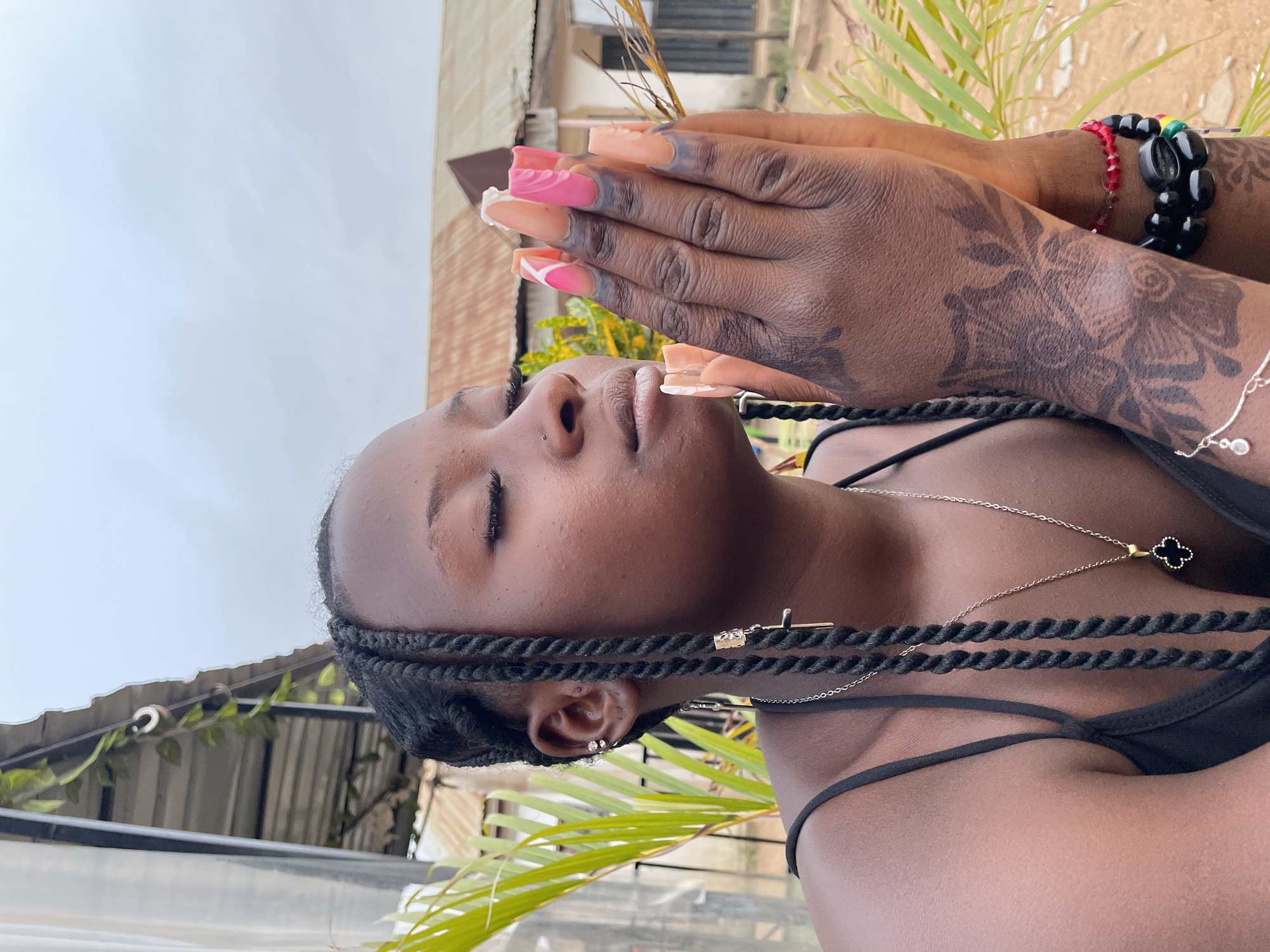A young, dark-skinned woman is shown in the image. She has her eyes closed, seemingly in a meditative or relaxed state. Her hair is styled in braids, and she wears a black top. Several pieces of jewelry are visible: a silver necklace with a pendant, small earrings, and decorative items in her hair. Her hands are clasped together, positioned near her face, with long, stylized nails painted in pink and orange designs. Her hands and arms feature intricate henna patterns. She is wearing a bracelet made of red and black beads on one wrist and a silver bracelet with dangling ornaments on the other wrist.
The background shows hints of an urban environment, possibly in Jos, Nigeria, including the presence of buildings and greenery. The setting suggests an outdoor location, with natural light illuminating the scene.