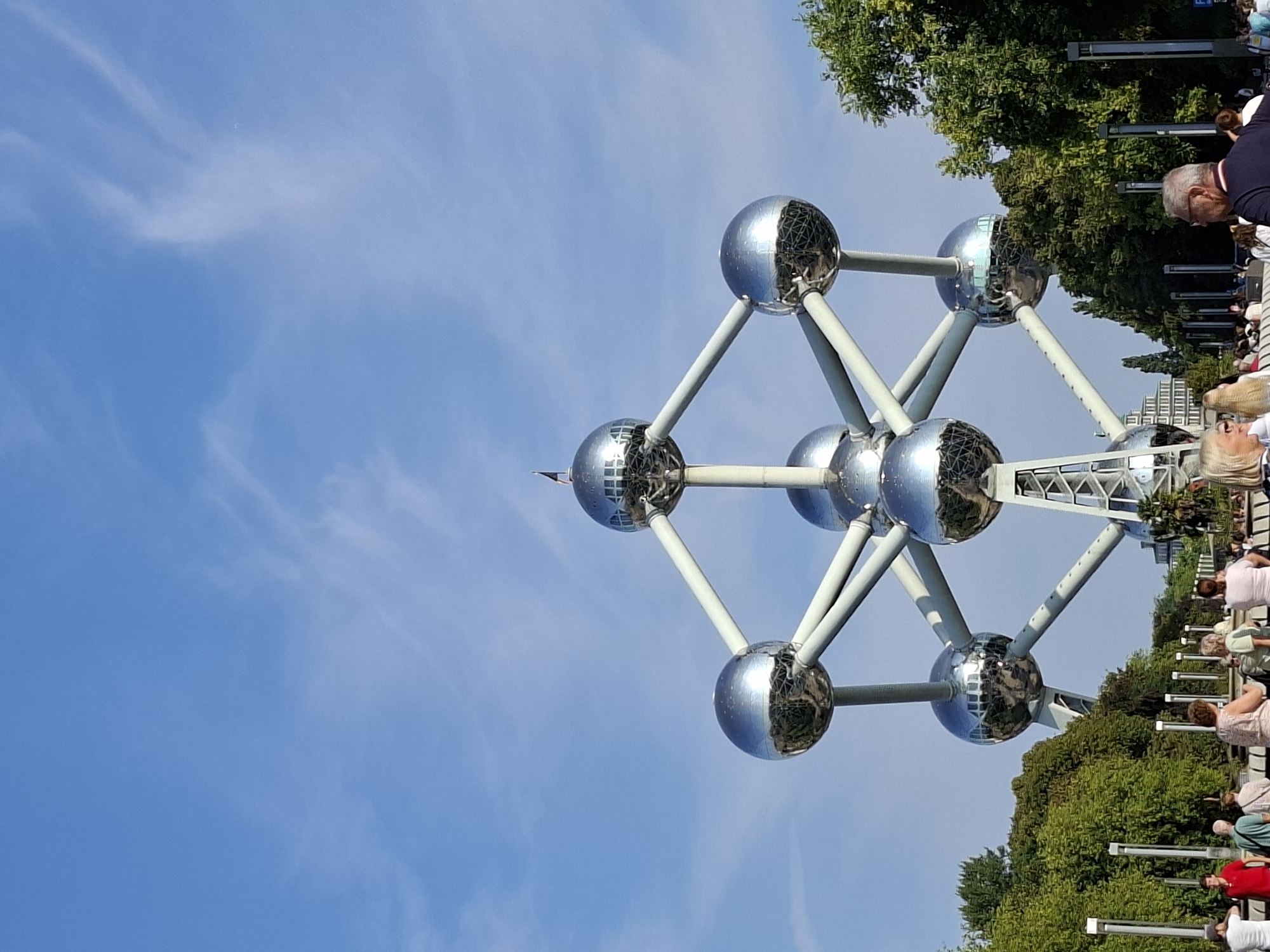 The image captures the Atomium, a landmark in Brussels, Belgium, under a bright blue sky with wispy white clouds. The iconic structure, resembling an enlarged iron crystal lattice, dominates the center of the frame. It consists of nine interconnected spheres, six of which are visible in this shot, all with a polished, metallic finish that reflects the surrounding environment. White struts connect the spheres, forming geometric patterns. A small flag is perched atop one of the upper spheres.
At the base of the Atomium, a crowd of people is visible, suggesting a public gathering or tourist activity. They are casually dressed and appear to be strolling or observing the monument. Trees with lush green foliage line the pathway in front of the Atomium, with some taller trees in the background and on the right side of the image. In the distance, beyond the trees, a multi-story building with a distinct architectural style is partially visible. The overall scene depicts a sunny day at a popular tourist attraction in Brussels.