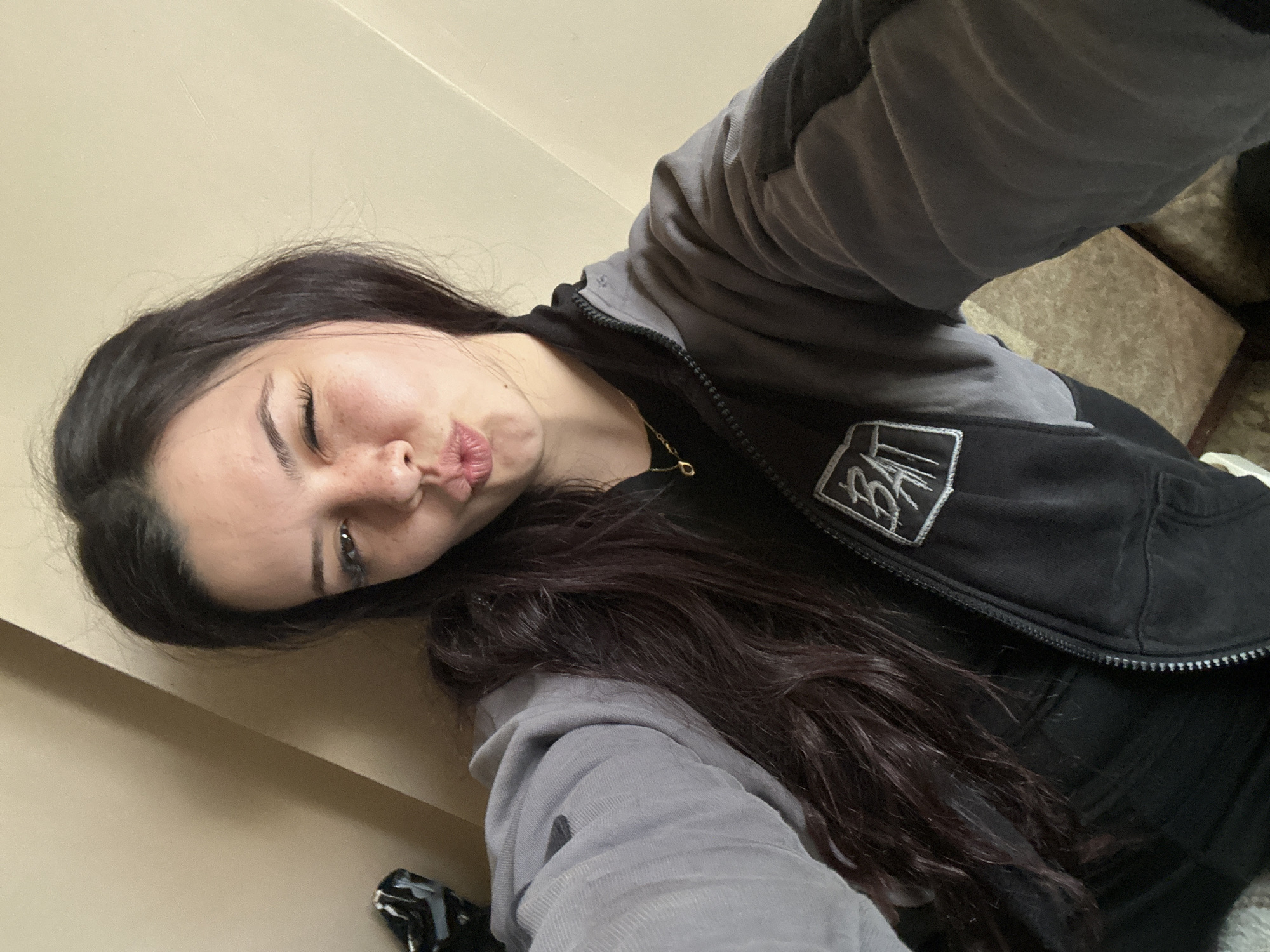 A woman is posing for a selfie indoors. She is angled, with her head tilted to the left. She has long, dark hair and is wearing a black and grey hoodie with a logo on the chest. She is making a kissing face towards the camera. The background consists of a light-colored wall and a glimpse of a patterned chair or cushion. There is nothing in the image to indicate the location in the city of Sliven, Bulgaria.