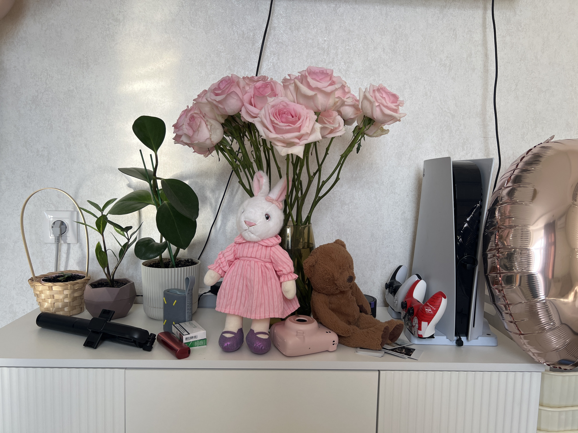 The image displays a collection of items arranged on a white cabinet against a textured, light-colored wall. A large bouquet of pink roses in a vase is centered in the composition. To the left, two potted plants and a wicker basket containing another plant are visible, near a white power outlet with a plug.
In front of the roses, a white stuffed bunny wearing a pink striped dress and purple glitter shoes sits, next to a brown teddy bear. A pink instant camera is positioned between the two stuffed animals. To the right, a white PlayStation 5 console stands vertically, flanked by two PlayStation controllers: one black and white, and another red and white with the partial text "CHA-CHA". A large, rose gold or bronze foil balloon is partially visible on the far right.
On the left side of the cabinet, a black selfie stick lies horizontally, alongside a red cylindrical object and a pack of HEETS cigarettes. A gray portable speaker is also present in this area. The scene is illuminated by soft, natural light, suggesting it is daytime. The arrangement creates a personal and decorative display, likely in a home setting such as a bedroom or living area.
