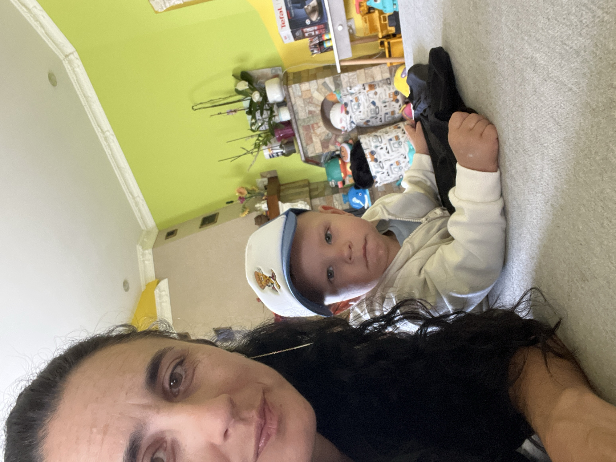 The image depicts a selfie-style photograph taken indoors, likely within a residential setting in Onokivtsi, Ukraine. In the foreground, a woman with long, dark, wavy hair is partially visible on the left side of the frame, her face turned towards the camera. To her right and slightly further back, a young child is lying down, also facing the camera. The child is wearing a light-colored hooded jacket and a white baseball cap with a cartoon image on the front. The child's gaze is directed forward, and their right hand is resting on a black piece of fabric.
The background reveals a room with lime green walls and a cream-colored ceiling. A fireplace with a stone facade is visible, decorated with plants in pots and other decorative items. To the right of the fireplace, a shelf unit holds various objects, including a "Tefal" branded appliance. Several toy storage bins, adorned with playful patterns of construction vehicles, are scattered around the floor. The overall environment suggests a family living space, possibly a living room or playroom, with toys and everyday household items present. The lighting appears to be a mix of natural and artificial light.