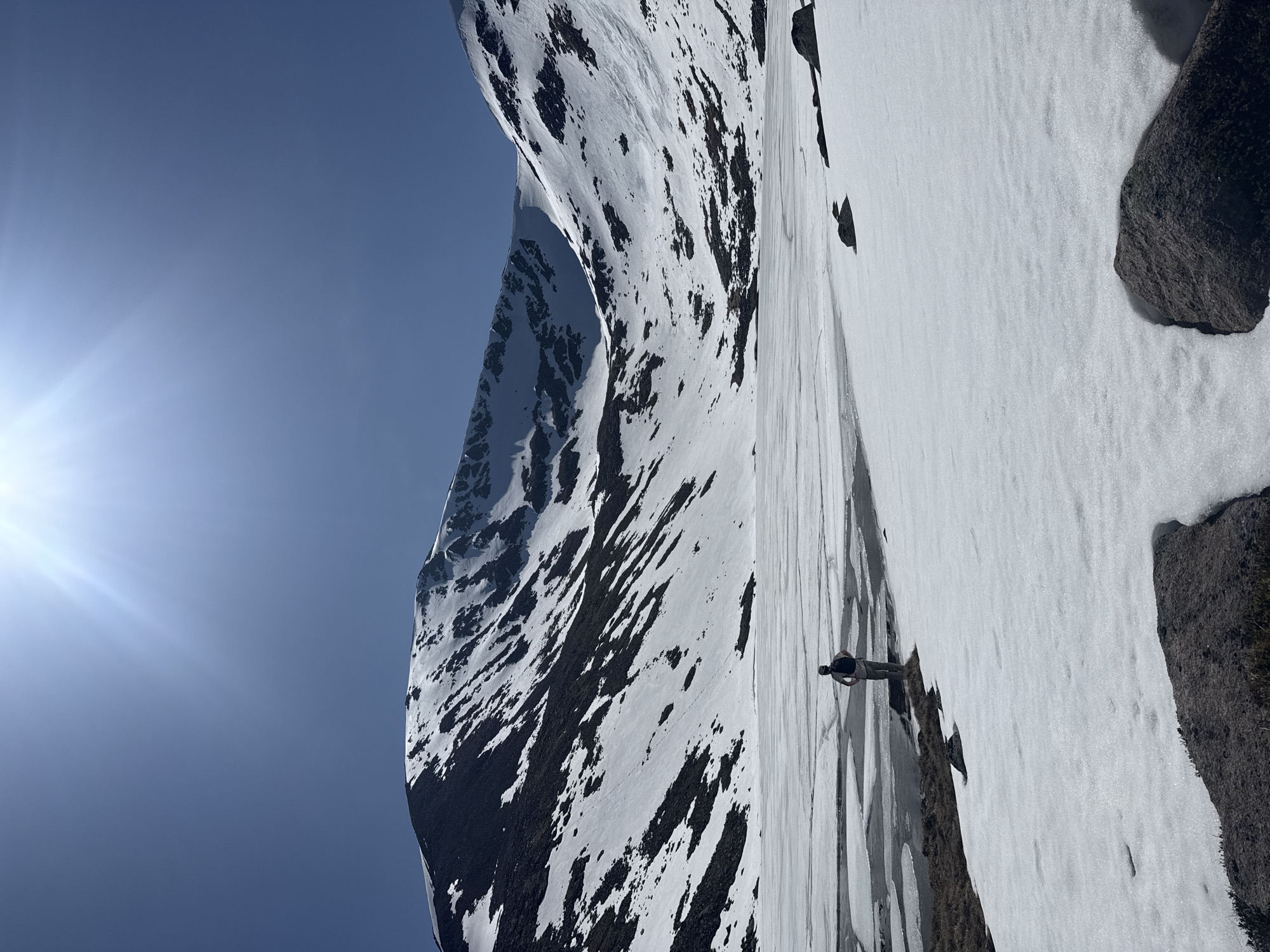 The image shows a snow-covered mountain landscape under a clear blue sky with a bright sun. The mountain features a mix of snow and dark rocky patches. A person stands on the snow, casting a shadow. Rocks are visible in the foreground.