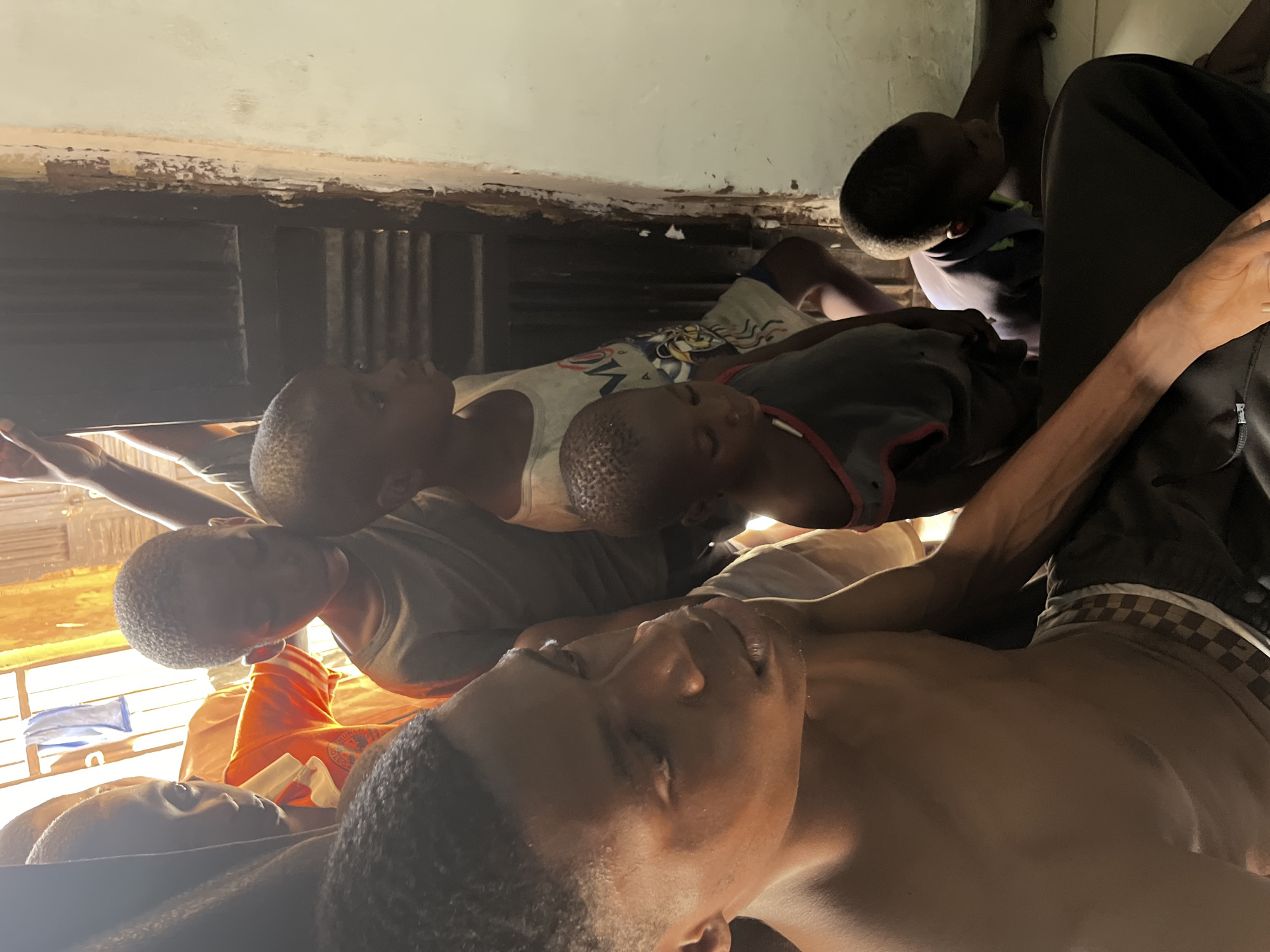 The image shows a group of young people, likely children or adolescents, lying down in what appears to be a dimly lit room. The subjects are lying close together, some on their backs and others on their sides, with their eyes closed, suggesting they are asleep or resting. One individual in the foreground is lying on their back, with their face clearly visible, appearing to be in a deep sleep. Another person is lying next to them, also with eyes closed. Further back, more individuals can be seen in similar positions, with their heads resting on the ground or on pillows.
The setting seems to be a simple, perhaps impoverished, living space. The walls are a pale, off-white color, with visible signs of wear and tear, including peeling paint and some dark staining along the top edge. There is a dark, slatted structure behind some of the subjects, possibly a window or a ventilation grate. The lighting is dim, with a warm, yellowish glow emanating from the right side of the frame, hinting at a light source outside or a window in that direction.
The overall mood of the image is one of quiet repose and vulnerability. The close proximity of the individuals suggests a communal living arrangement, and their peaceful sleep evokes a sense of shared experience or hardship. There is no discernible text in the image. The time of day is unclear, but the subdued lighting could suggest either early morning or late evening.