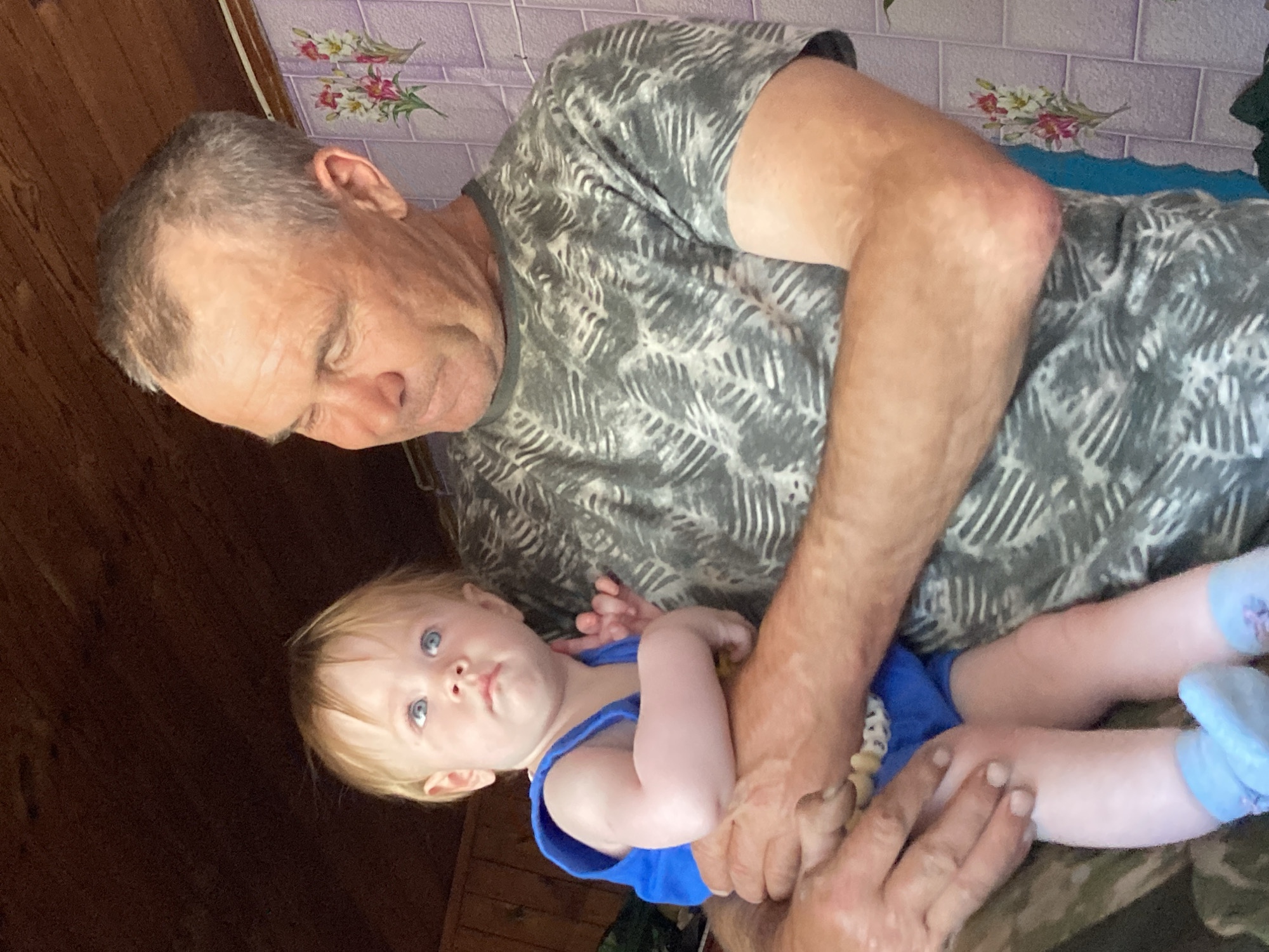 In an indoor setting, a Caucasian man with short gray hair and weathered skin is holding a fair-skinned baby. The man wears a gray t-shirt with a white palm-leaf pattern and appears to be looking down at the baby. The baby has fair hair and blue eyes and is looking upwards. The baby is wearing a blue sleeveless top and blue booties. The background consists of dark wooden paneling on the ceiling and a tiled wall with a floral pattern. The scene is likely located in the village of Rakity, Russia, given the provided context.