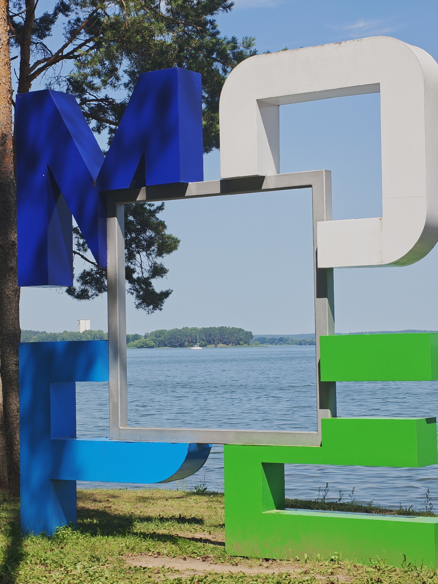A large, colorful metal sculpture is positioned in front of a body of water and a treeline under a clear blue sky. The sculpture is composed of several geometric shapes, predominantly in the colors blue, white, and green. The letters "M" in blue, a large white arch, and green rectangular forms are visible, suggesting it may spell out a word or phrase. A silver metal frame is integrated into the sculpture, framing a view of the water, distant trees, and a small white sailboat on the water. The foreground shows a grassy area with some scattered pinecones and dried leaves, indicating an outdoor park or waterfront setting. The presence of a tall, light-colored building is faintly visible in the distance, beyond the treeline on the far side of the water. The overall scene is bright and sunny, suggesting a summer day. The location is in Ждановіцкі сельскі Савет, Belarus.