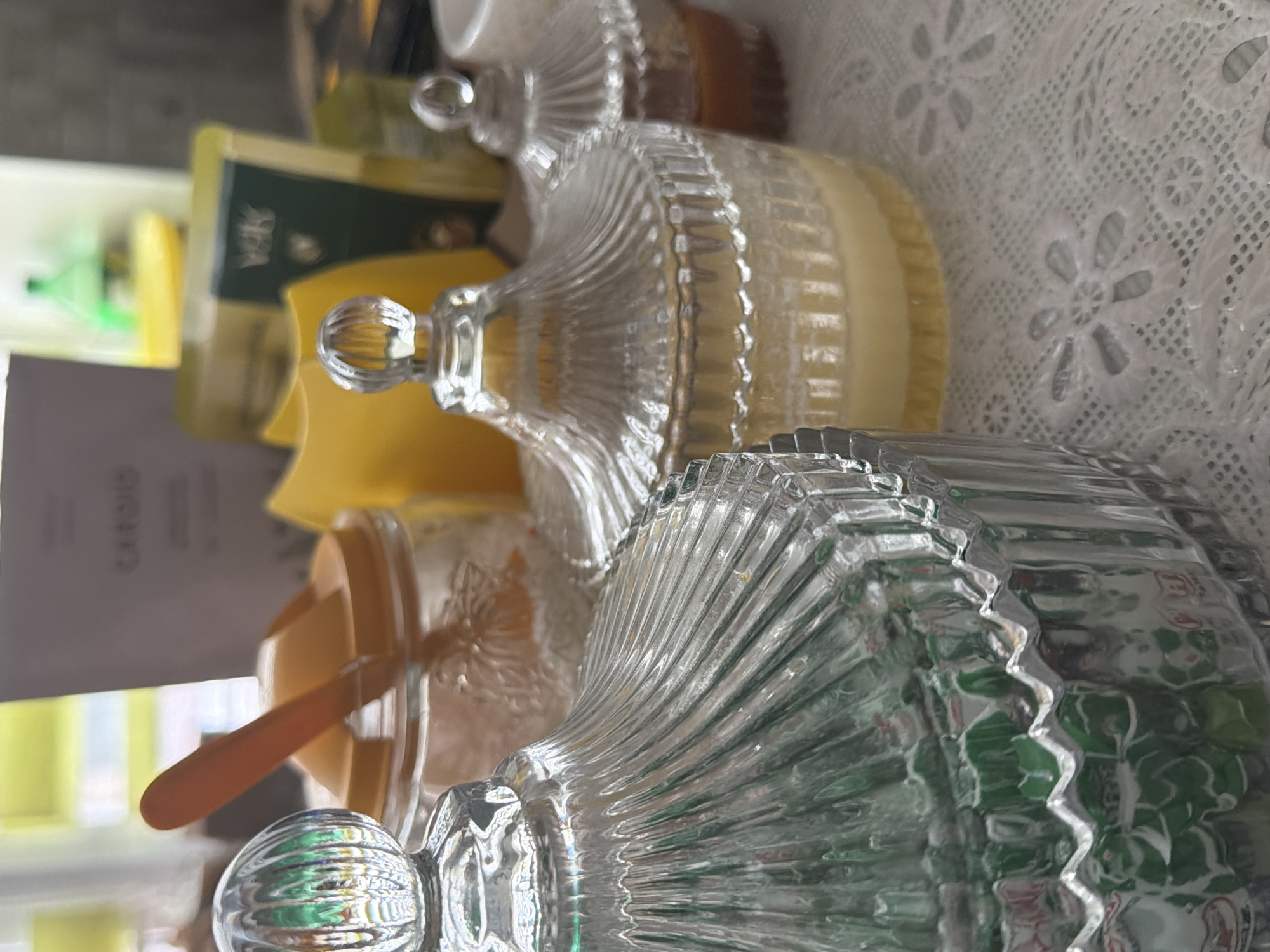 The image shows a close-up of two ornate, clear glass jars with lids, seemingly resting on a white, lace-patterned surface. The jars are partially filled with substances, one appearing yellowish-white and the other greenish. A wooden spoon-like object is visible within one of the jars. The background includes blurry shapes of boxes and other containers. The image quality is soft and partially out of focus. No people, specific actions, or identifiable location within Chaikovsky, Russia, are evident.