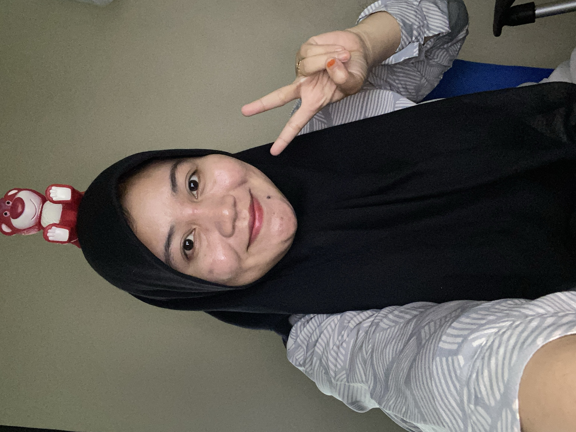 A young woman wearing a black hijab and a grey and white striped shirt is taking a selfie. A red plush toy is balanced on top of her head. She is smiling and holding up two fingers in a peace sign. Her right hand has orange nail polish on one fingernail and a ring on her ring finger. She appears to be indoors, with a neutral-colored wall behind her. The scene is likely in Banjarbaru, Indonesia, though no specific landmarks are visible to confirm the location within the city.