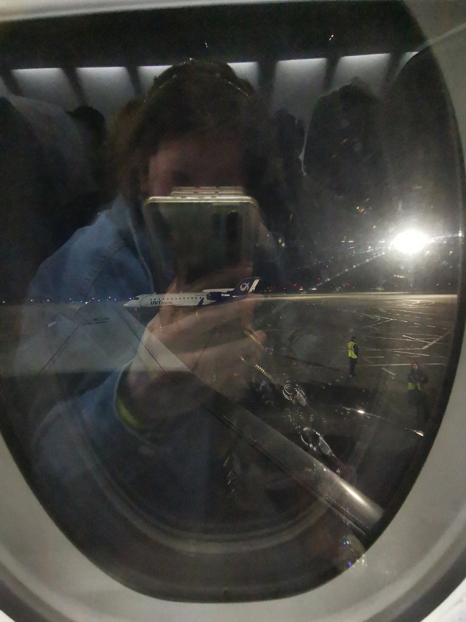 A blurry view from an airplane window shows the reflection of a person holding a smartphone to take a picture of an aircraft parked on the tarmac. The plane outside the window appears to have "UVT aero" printed on its side. Two workers in yellow vests are visible on the tarmac. The setting seems to be at night, with bright lights illuminating the scene. The city is Новое Мартьяново, Russia.
