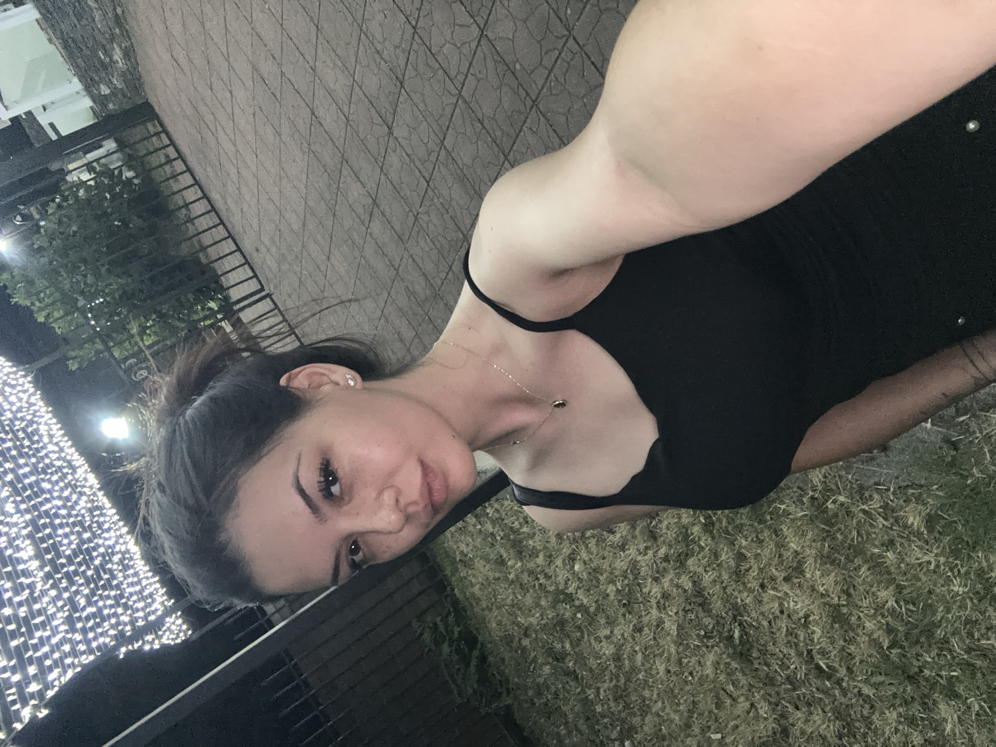 A young woman with dark hair pulled up in a bun takes a selfie outdoors at night. She is wearing a black tank top and a delicate necklace. She has freckles across her nose and cheeks and is wearing small, sparkly earrings. Behind her, there is a paved area with a stone pattern, a metal fence, and some greenery. Above, there is a canopy of string lights. The scene appears to be in an urban setting, possibly a park or outdoor venue, in Sliven, Bulgaria.