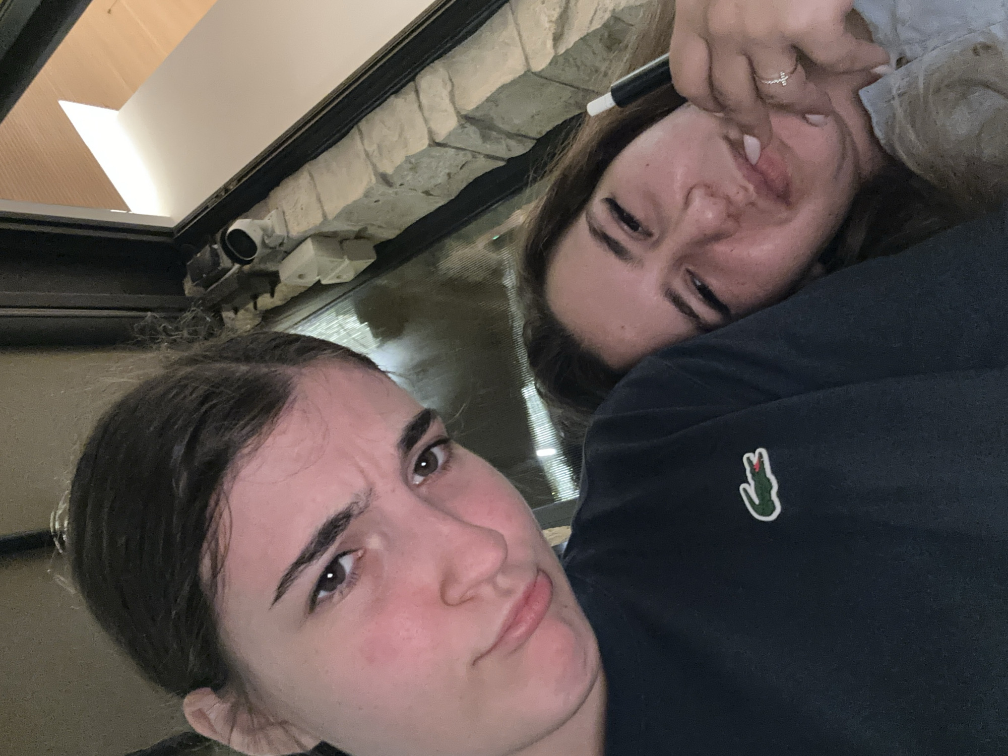Two individuals, presumably female, are captured in a close-up, high-angle selfie. The person in the foreground, on the left, has dark brown hair pulled back, revealing her face with a stern expression and downturned lips. She is wearing a dark blue t-shirt with a white and green Lacoste logo. The second individual, on the right, is partially visible, with long brown hair and a grimacing expression, her hand near her mouth. She appears to be wearing a light blue garment and a ring on her right ring finger.
The background suggests an indoor setting with a stone-like textured wall. A window or opening in the structure reveals an illuminated space with wooden elements, possibly indicating a modern interior design or architectural feature. A small security camera is visible mounted on the stone structure near the window. The lighting is dim, with the primary light source appearing to be from the illuminated area in the background. The overall scene appears to be casual and personal, likely taken within a residential or communal living space. The exact location in Nicosia, Cyprus, cannot be determined from the visual information provided.