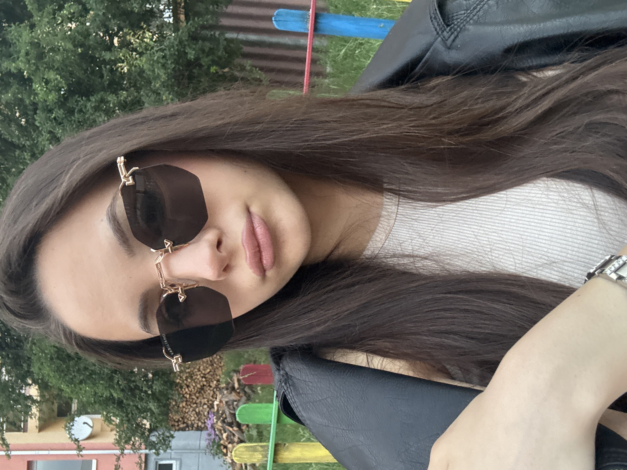 A front-facing photograph captures a young adult female outdoors in an unknown residential or common area. The subject, positioned centrally, has long, straight, dark brown hair falling over her shoulders and is wearing dark-tinted, frameless, hexagonal sunglasses with gold-tone hardware; the brand "VASILIANO" is visible on the left temple. Her lips are slightly pursed in a pout. She wears a light-colored, possibly ribbed, tank top or undershirt, partially obscured by a black faux leather jacket draped over her left shoulder (viewer's right). A silver-tone link bracelet watch is visible on her right wrist (viewer's left).
The background features a low, multi-colored wooden picket fence or barrier running horizontally through the mid-ground, displaying yellow, green, red, and blue painted sections. Behind this fence, on the left, is a neatly stacked pile of cut firewood and some small purple flowering plants. Further left, a multi-story building with peach/reddish-pink and lighter-colored sections is partially visible, featuring a white satellite dish mounted on its side and a white-framed window. Dense green foliage, indicative of trees or bushes, occupies the upper right and central left background. On the far right, behind the colorful fence, a section of corrugated brown or dark gray fencing is visible. Patches of green grass are present in the lower right background.