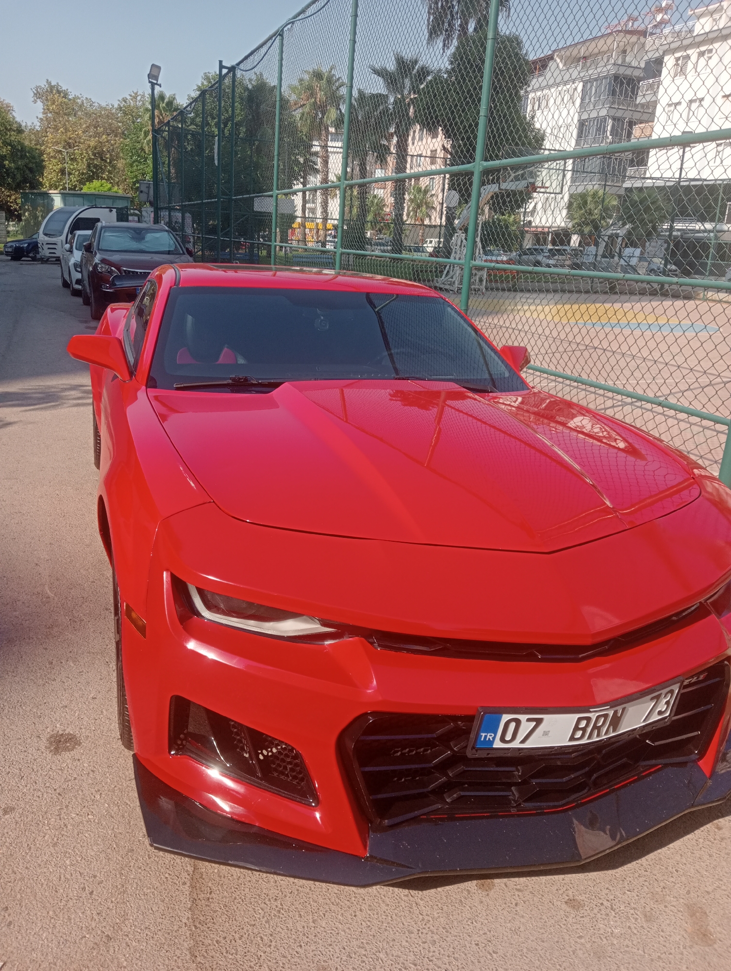 The image shows the front of a red Chevrolet Camaro parked on a paved street in Antalya, Turkey. The car's license plate reads "TR 07 BRN 73". In the background, a chain-link fence encloses a basketball court with visible court markings. Beyond the court are palm trees and several multi-story buildings. Other cars are parked in a line behind the Camaro. The scene appears to be a sunny day.