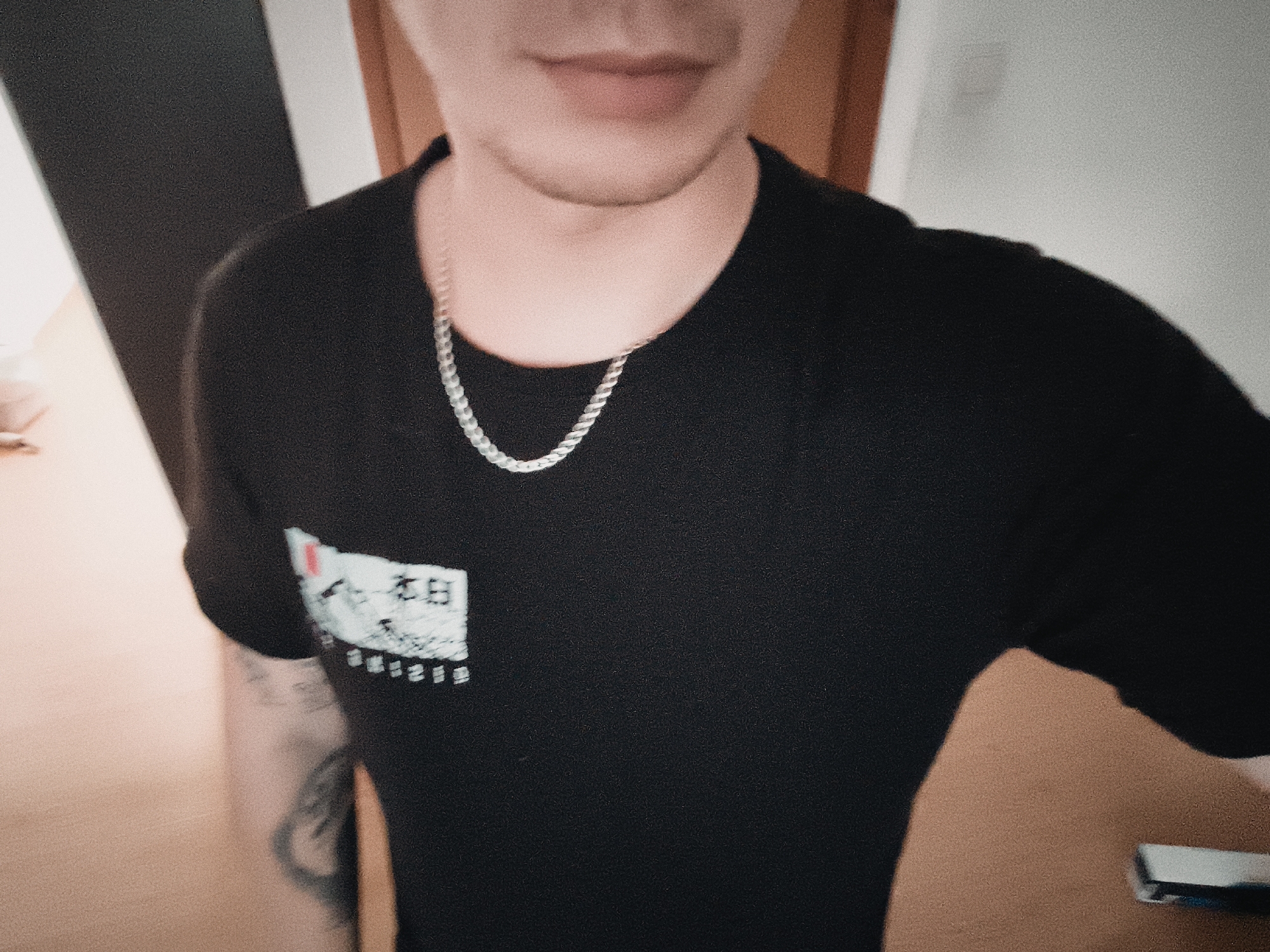 A person wearing a black t-shirt with a graphic and a silver necklace is shown in an indoor setting. The graphic on the t-shirt is white with black text. A tattoo is visible on the person's left arm. A wooden door and frame are in the background. The location is not discernible as being in Nowy Tomyśl, Poland, based on the visual information provided.