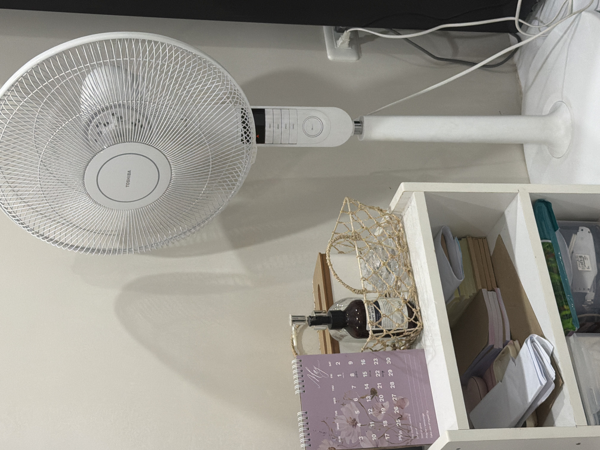 The image depicts a white Toshiba electric fan standing on a white shelf unit against a white wall. The fan has a circular grill and a control panel on its stem. Wires from the fan are visible near the top of the image, near a wall outlet. On the shelf beneath the fan, there's a decorative wire basket containing a dark glass bottle with a pump dispenser and packaging, along with a calendar displaying the month of May. Also within the shelf are stacks of papers and other miscellaneous items. The location appears to be indoors, but the specific details necessary to geolocate the image to Davao City, Philippines, are not present.