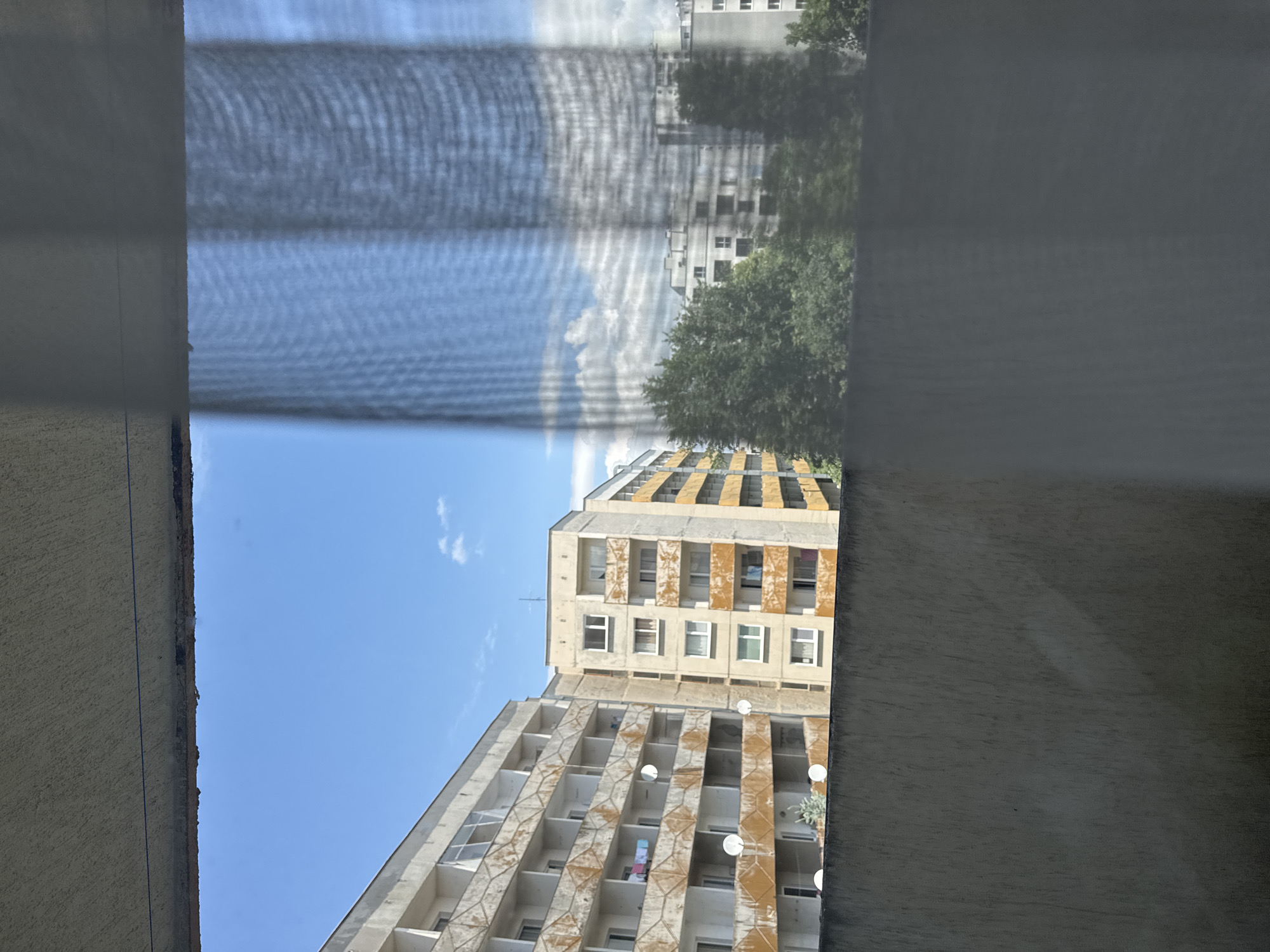 A forensic view captures an outdoor scene from a high vantage point, likely a balcony or window, in a residential area of Sofia, Bulgaria, identifiable by the characteristic post-socialist era concrete residential blocks. The image is bisected vertically by a semi-transparent dark grey mesh curtain that obscures the right half.
The upper section of the image features a rough, textured light grey concrete ceiling or overhang, with a thin, horizontally taut blue line extending across its underside. Below this, a clear blue sky with minimal scattered white clouds occupies the upper left quadrant.
The left side of the scene displays two prominent multi-story residential buildings under bright daylight. The building on the far left is a large, weathered concrete structure with numerous open balconies featuring distinctive geometric patterns on their balustrades. These balconies exhibit significant orange-brown discoloration and streaking, suggesting rust or aged paint. Several white circular satellite dishes are affixed to various balconies. Visible laundry hangs on a balcony near the middle of this building. Behind it and slightly to the right, a second residential building, appearing a lighter yellowish-beige, stands with white-framed windows and similar, though less geometrically patterned, discolored balconies.
The right half of the image is viewed through the obscuring mesh curtain, which creates a visible grid pattern. Through this curtain, a lighter blue sky with larger, more defined white clouds is discernible. In the mid-ground, a dense cluster of green trees partially obscures other distant, multi-story residential buildings, which appear less distinct due to both distance and the curtain's interference.
The lower third of the image is dominated by a dark grey, textured concrete wall or parapet, likely belonging to the vantage point, showing subtle variations in tone and texture. No people or overt actions are visible.