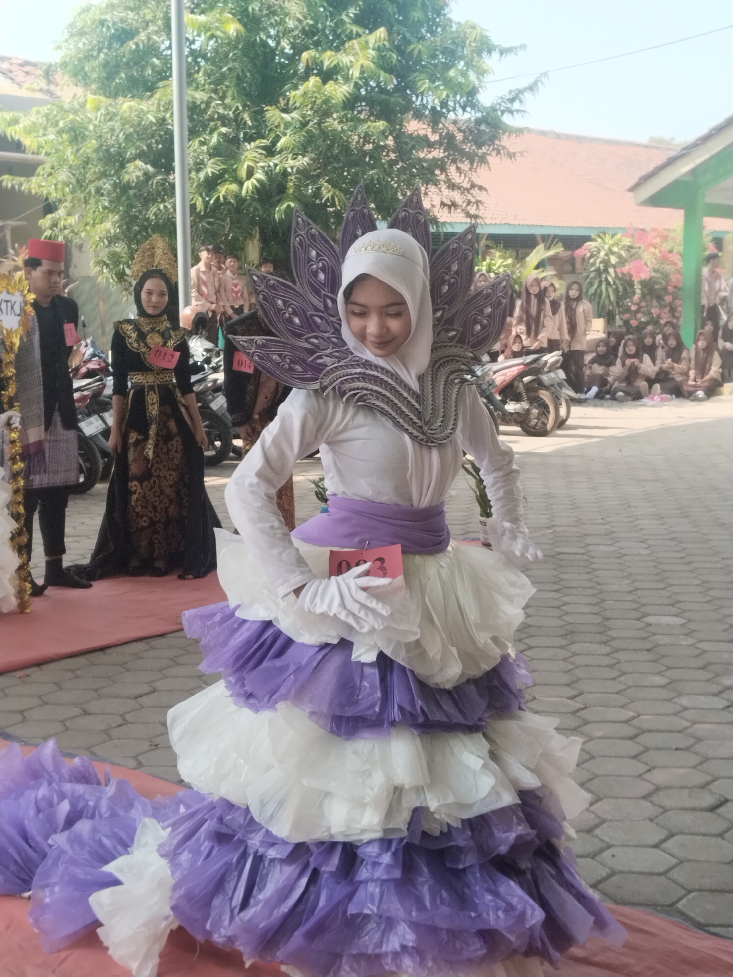 In Pakulaut, Indonesia, a young woman wearing a white hijab and a white shirt, adorned with a voluminous, multi-layered dress of white and purple ruffled fabric, participates in an event outdoors. The dress features an elaborate, purple, lotus-like collar embellished with intricate patterns and what appear to be small, decorative gems. A purple sash cinches her waist, and she wears white gloves. A pink tag with the number "03" is visible on her dress. To her left, a woman in a black and gold traditional gown with a golden headpiece, identified by a pink tag with "012," stands on a red carpet. Behind them, other participants and spectators, some wearing scout uniforms, are gathered. Motorcycles are parked in the background. The scene takes place on a paved area with hexagonal tiles, adjacent to a building with an orange-tiled roof and a green structure. Trees with lush green foliage are prominent in the background.