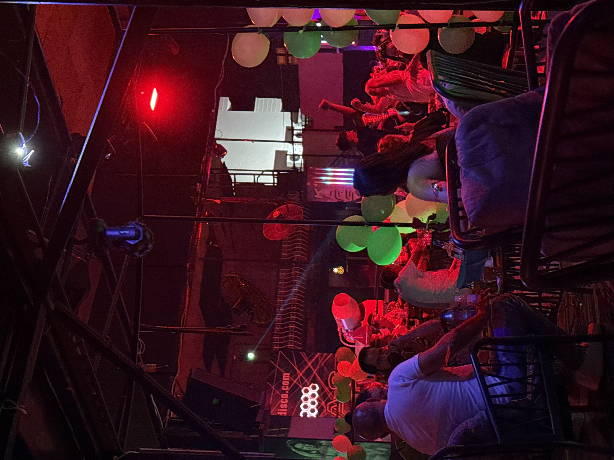 The image depicts the interior of a disco club in Alanya, Turkey. The environment is dimly lit with red and purple lighting, characteristic of a nightlife venue. In the foreground, several individuals are seated at tables, some are seen from behind, while others are partially visible. They appear to be patrons of the club, engaged in conversation or observing the scene. Prominently displayed are numerous balloons in various colors such as green, yellow, and red, adding a festive atmosphere. The background reveals a stage area with a large screen displaying "disco.com" and geometric patterns, suggesting entertainment or a promotional display. Overhead, industrial lighting fixtures and structural elements of the building are visible. The overall impression is of a lively and vibrant disco club scene in Alanya.