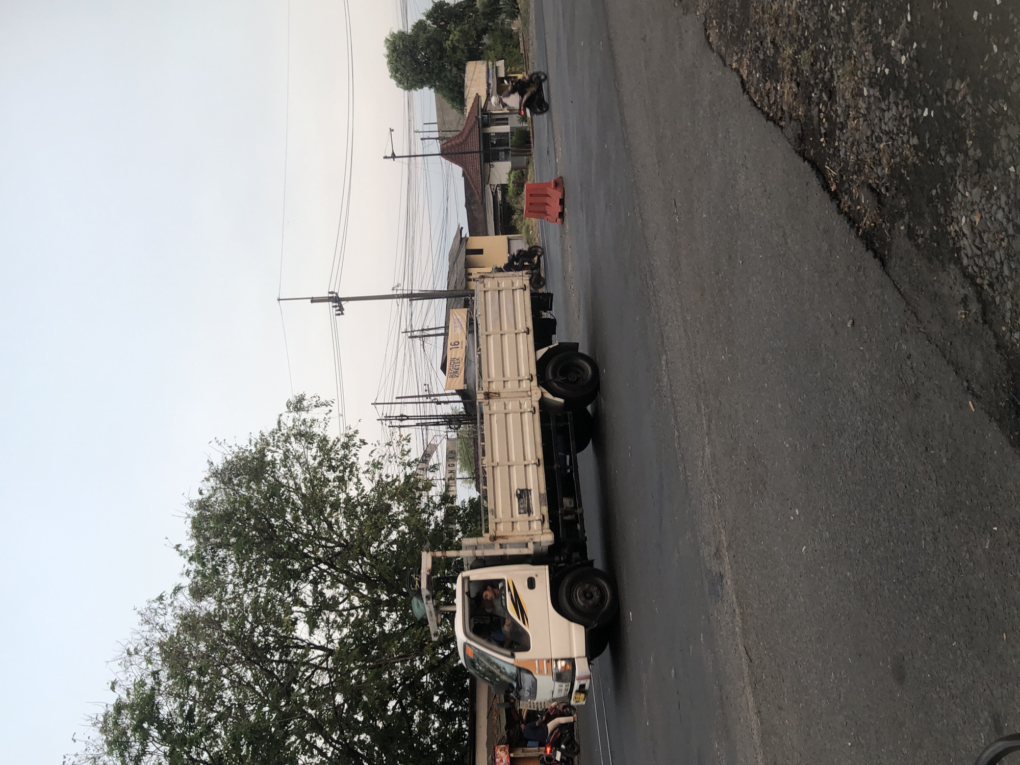 A white flatbed truck is traveling on a road in Siring, Indonesia. The truck has a beige colored cargo bed with vertical metal slats. The truck's cabin is white with yellow and black accents on the side. A driver is visible in the cabin. In the background, there are buildings, trees, power lines, and other vehicles including motorcycles. A red traffic barrier is visible on the right side of the road. The sky is overcast and light.