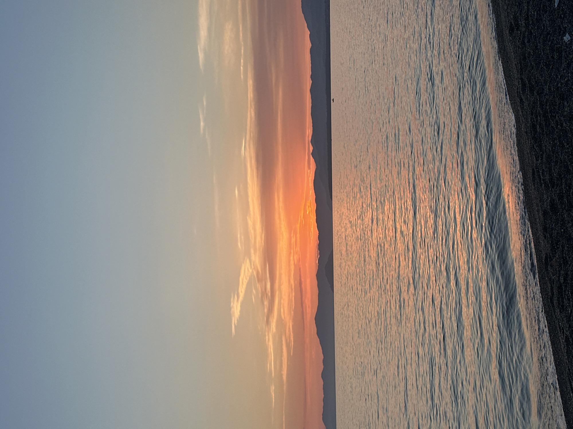 The image depicts a serene sunset over the sea, with a silhouette of mountains in the background. The sky transitions from a pale blue at the top to vibrant shades of orange and pink near the horizon, where the sun is setting behind the mountains. Wispy clouds are scattered across the sky. The water in the foreground is calm, reflecting the colors of the sunset, with gentle waves lapping at a pebbly shore. In the distance, a small, dark silhouette of a boat can be seen on the water. This scene is consistent with the coastal landscape found in areas surrounding Fethiye, Turkey, known for its beautiful sunsets and mountainous coastline.