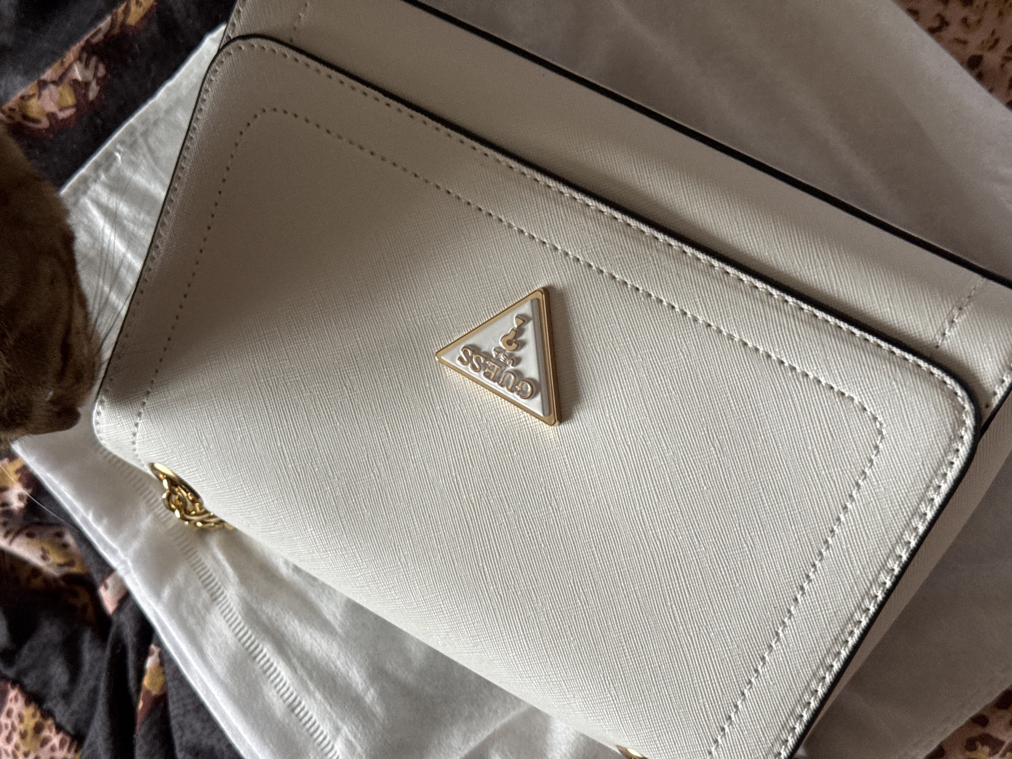 The image shows a close-up of a white Guess crossbody bag. The bag has a textured Saffiano leather finish and a gold-toned Guess logo in a triangular shape. The logo is embossed with the brand name and "USA". The bag is slightly open, revealing a glimpse of its interior. A gold chain strap is visible, attached to the side of the bag. In the background, a cat's head and paw can be seen to the left of the bag, with its fur appearing to be reddish-brown. The bag is placed on a white fabric, possibly a dust bag, which is lying on a patterned surface, possibly a blanket or rug with a leopard-like print. The lighting suggests it is daytime, possibly indoors, as there are no strong shadows or indications of outdoor weather. The overall impression is one of a new item being presented or examined. There is no discernible information about the specific time of day, weather, or emotional context beyond the presentation of the object. The location context of Izhevsk, Russia, is provided but not evident from the visual cues in the image.