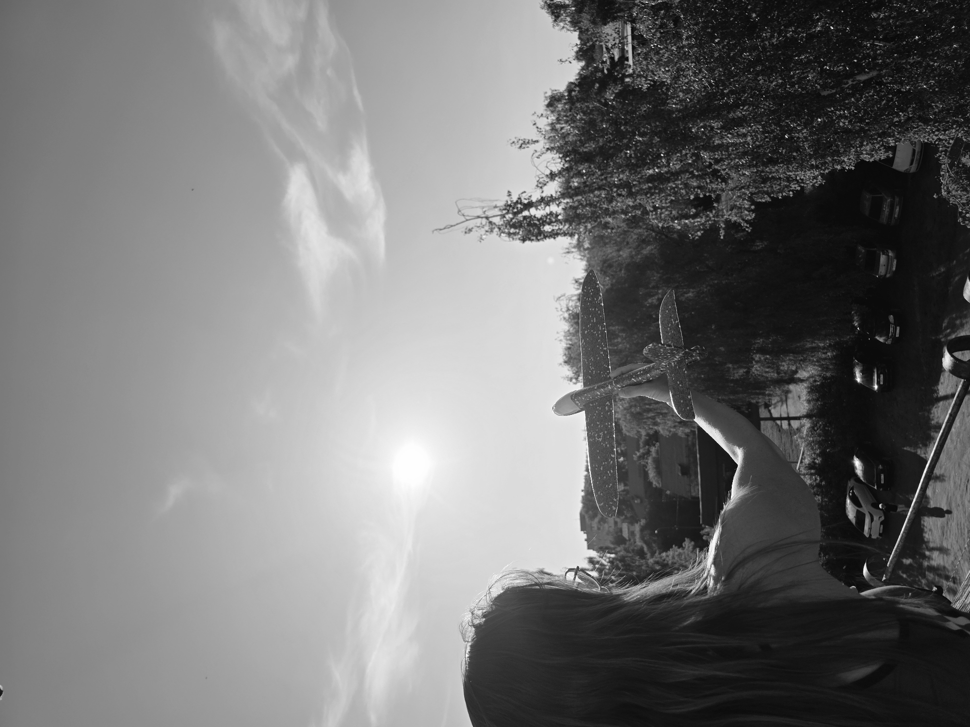 A black and white photograph, rotated 90 degrees counterclockwise, depicts a person holding a toy airplane towards the sky. The person, seen from behind with long hair, is holding the airplane with their right arm extended. The sun is positioned in the upper-left quadrant, creating a strong backlighting effect. The background features trees and buildings, with what appears to be parked cars on a road adjacent to the treeline. The location is Chaikovsky, Russia.