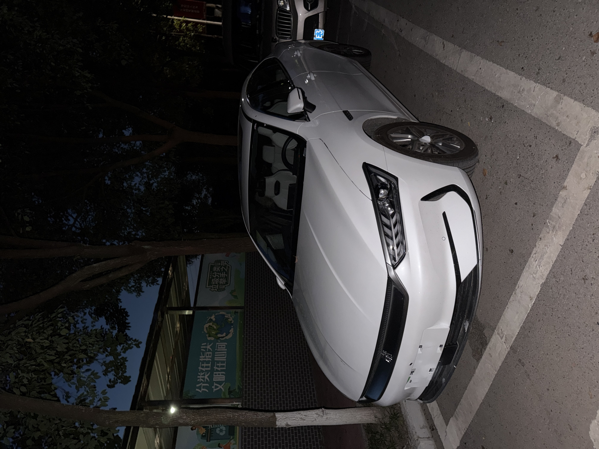 In the city of Jiangjin, China, a white BYD Han EV sedan is parked at night on a paved surface marked with white parking lines. The car is positioned at an angle, facing slightly to the right. The front of the car is prominently displayed, showcasing its sleek design, including headlights and a black grille with a logo. The interior of the car is visible, with light-colored seats and a steering wheel. The background features trees with dark foliage, suggesting an outdoor environment. A portion of another vehicle, a dark-colored SUV with a license plate displaying "E3", is visible to the right of the BYD. In the background, behind the car, there are banners displayed on a brick wall. One banner, in Chinese characters, reads "分类在指尖 文明在心间," which translates to "Classification at your fingertips, civilization in your heart." Another banner, partially obscured, also appears to be related to recycling or environmental awareness. The lighting suggests it is evening or night, with artificial light illuminating the scene. There are no people visibly present in the image.