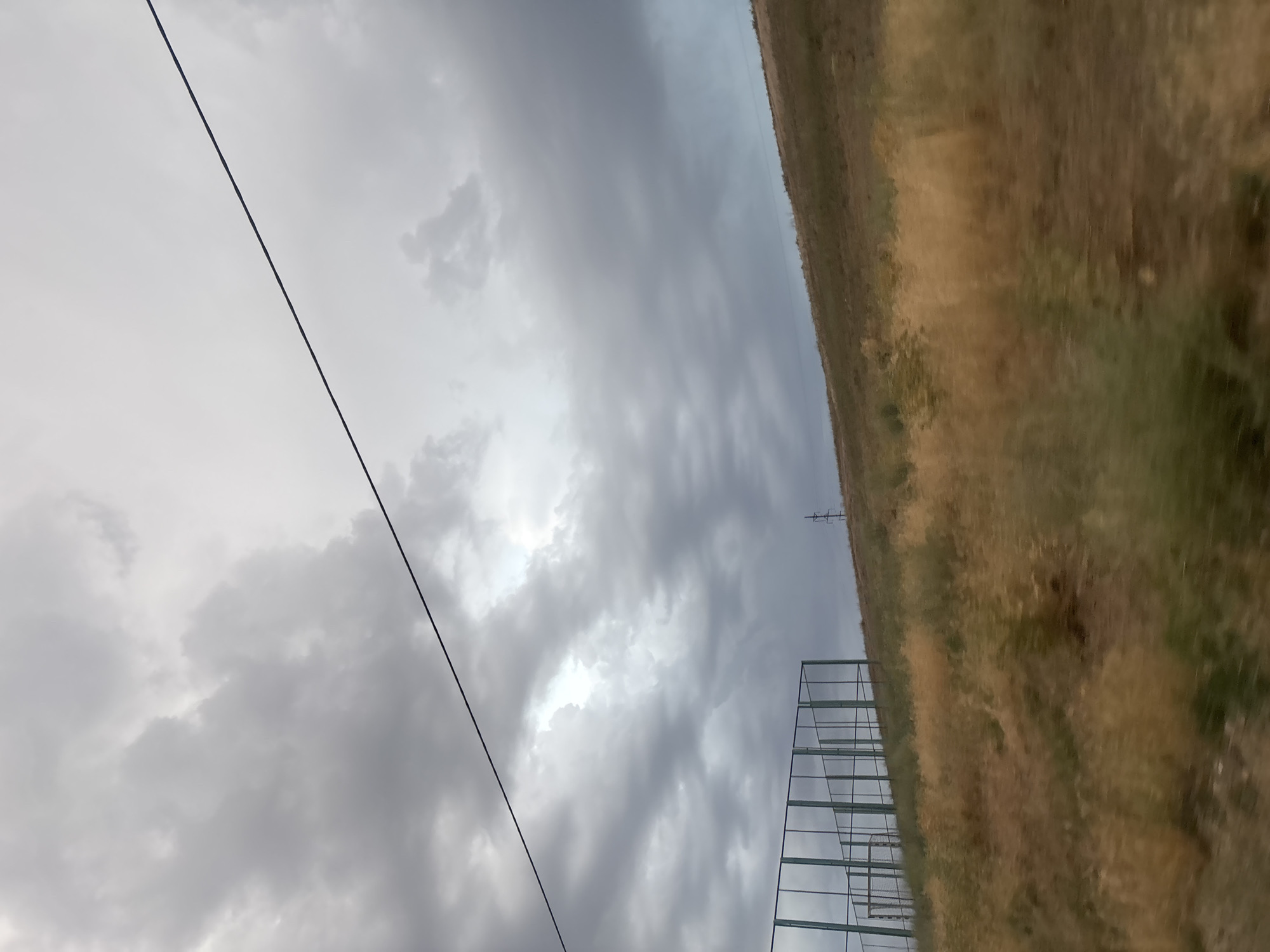 A cloudy sky looms over a grassy hill in Taraz, Kazakhstan. A wire stretches diagonally across the frame from the upper left to the upper right. In the lower left, a green chain-link fence encloses what appears to be a sports field, with a soccer goal visible. The hill is covered in dry, yellow grass. A solitary utility pole stands on the top of the hill in the middle ground.