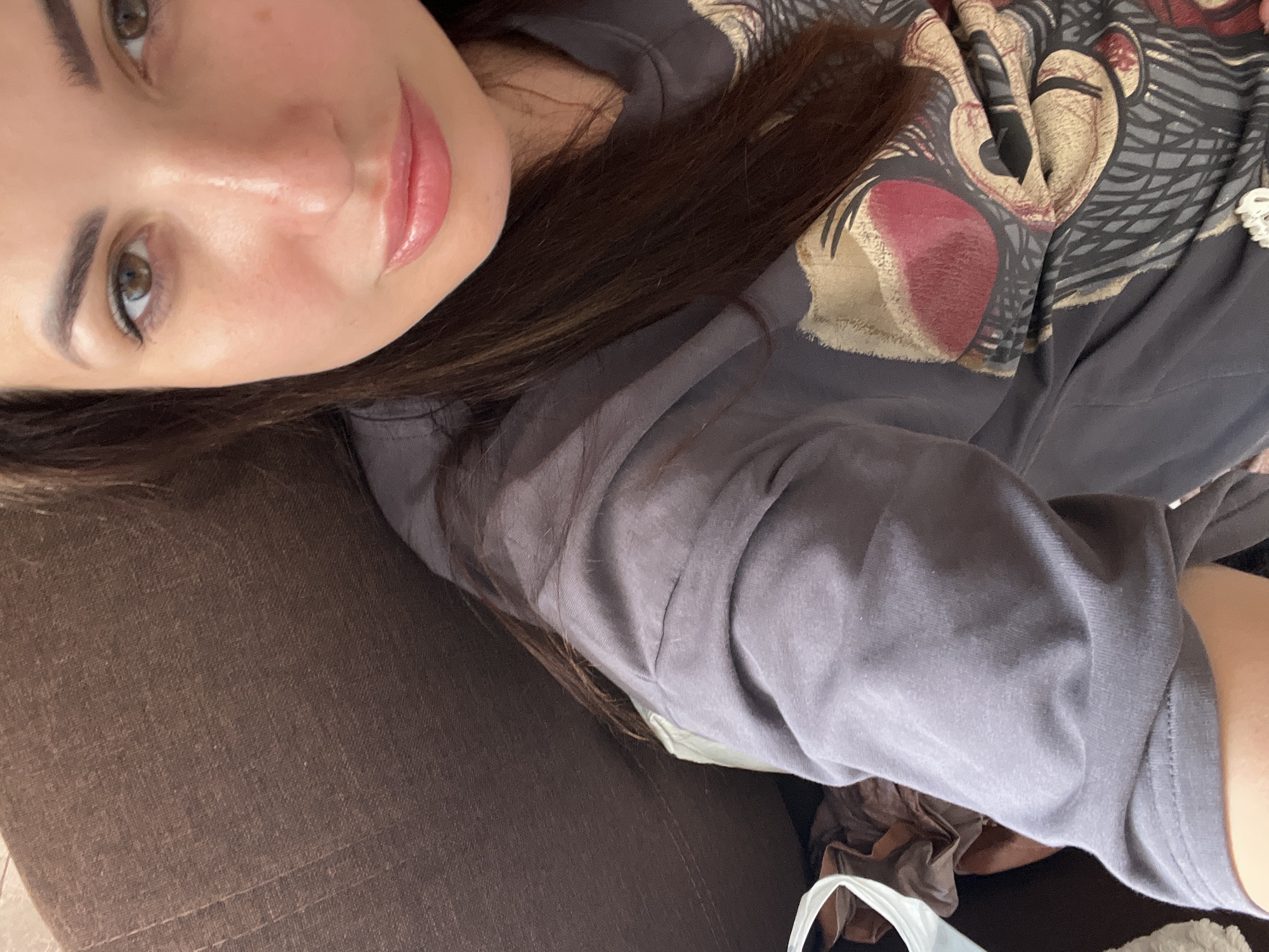 A woman with brown hair and brown eyes is captured in a close-up shot, wearing a gray t-shirt with a graphic print. She is seated on a brown couch, and her face is turned towards the camera. The image is focused on her upper body and face, with the couch occupying the left side of the frame. The lighting appears natural, casting a soft glow on her skin, and her expression is neutral.