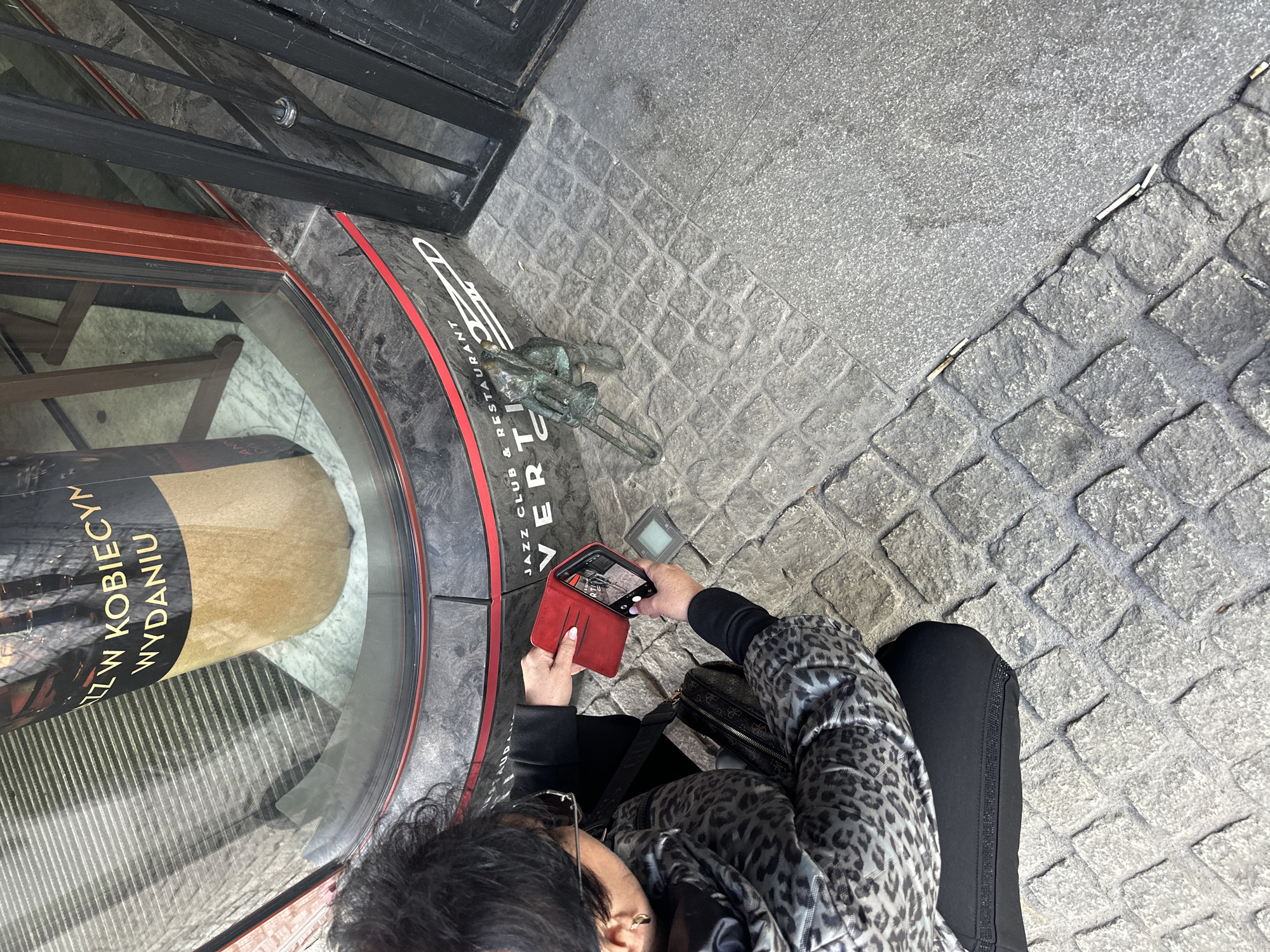 An overhead shot shows a person taking a picture with a smartphone. The person is wearing a leopard-print jacket and black pants and is situated on a cobblestone street next to a restaurant or jazz club called "Vertigo," located in Wrocław, Poland. A bronze sculpture of a musician with a trombone is affixed to the ground. The person is holding a red phone case open and using the camera app on the phone. A purse is nearby. The scene is outdoors, with grey cobblestones and adjacent surfaces visible.