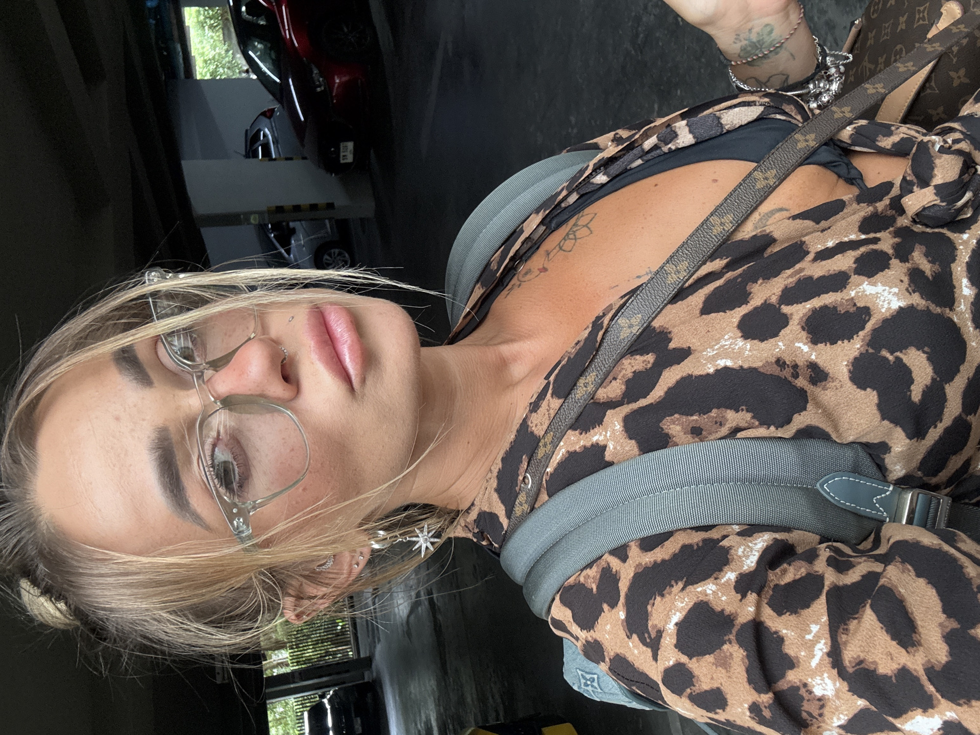 The image shows a close-up, selfie-style shot of a woman wearing clear-rimmed glasses, a leopard-print top, and a black bikini top. She has blonde hair pulled up in a messy bun and is wearing hoop earrings with a star charm. A grey backpack is visible over her shoulders, and a designer cross-body bag is draped across her chest. Her left arm is resting on the backpack strap, and her right arm is out of frame, but a bracelet and a tattoo are visible on her wrist.
The setting appears to be an indoor parking garage, likely in the city of Choeng Thale, Thailand, based on the presence of a dark-colored car and a red car parked in the background, as well as the lush green foliage visible through an opening in the garage structure. The lighting is dim and overcast, suggesting an indoor or shaded outdoor environment. The woman's expression is neutral, and she is looking directly at the camera.