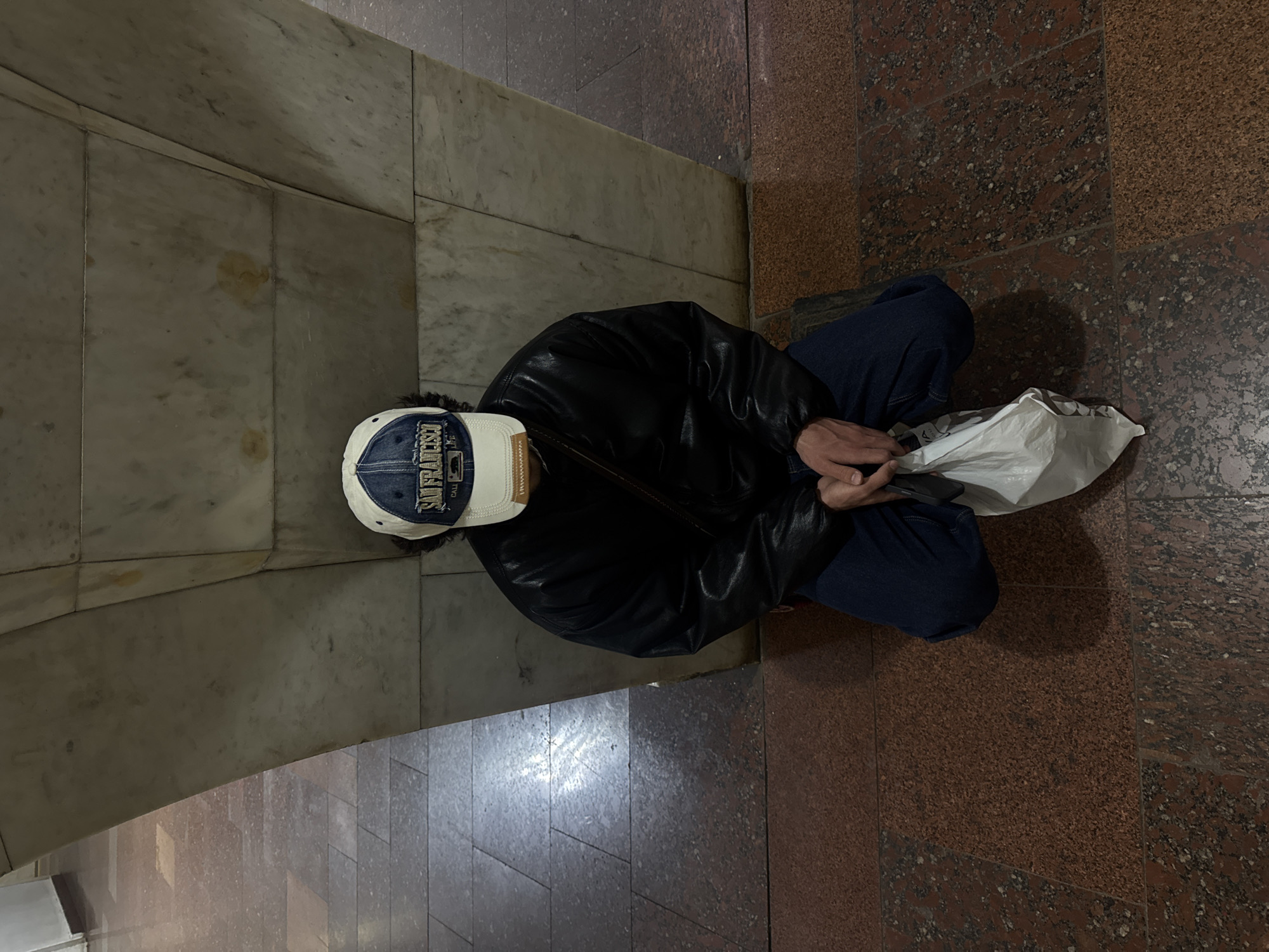 A top-down view shows a person seated in what appears to be the Yerevan Metro, Armenia. The person is wearing a dark leather jacket, blue jeans, and a baseball cap with "San Francisco" stitched on the front. They are holding a crumpled white plastic bag. They are seated against a wall with a curved corner, clad in light-colored marble-like panels. The floor is red and brown granite tiles of varying sizes. The person appears to be resting.