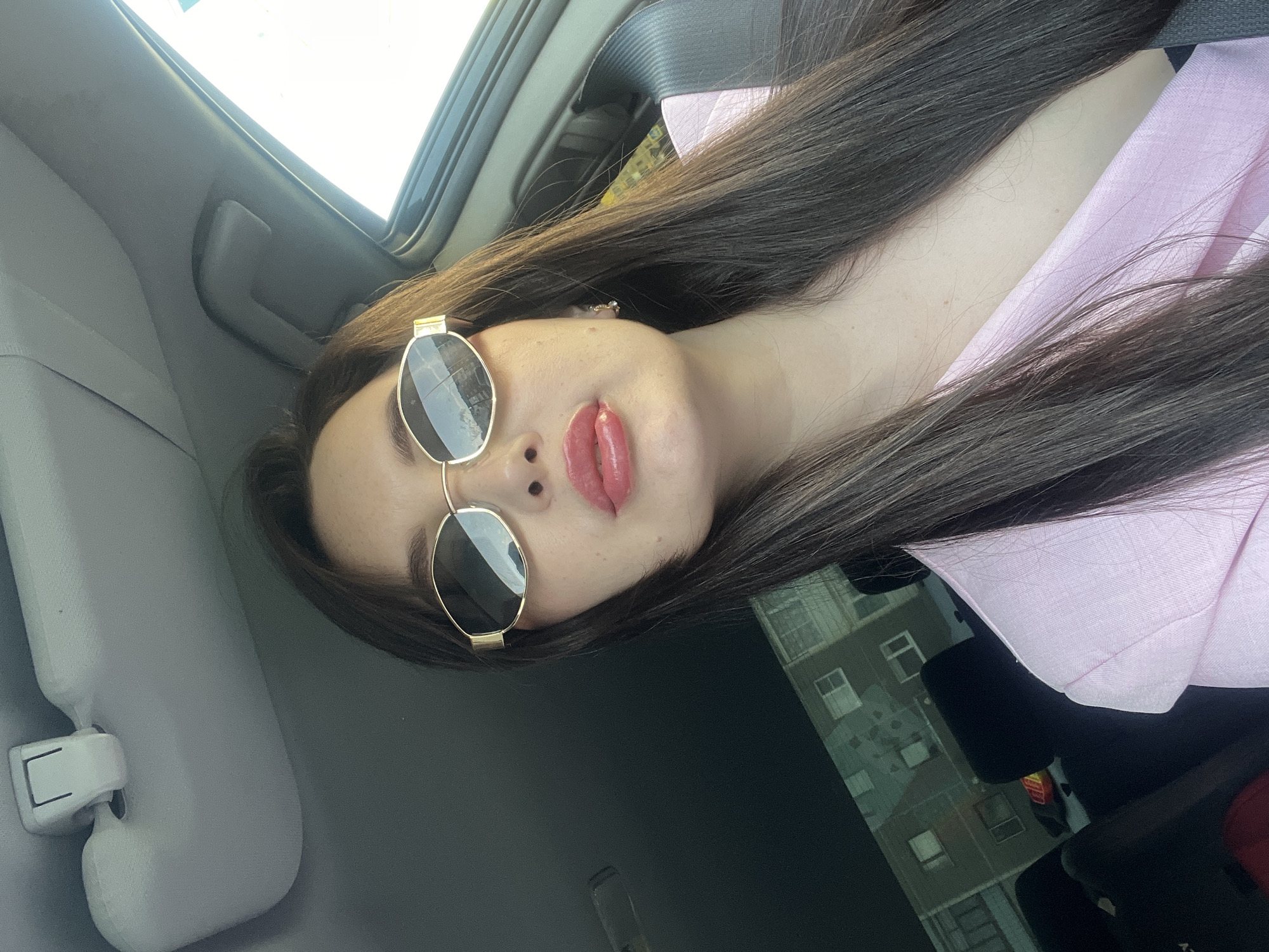 A woman is sitting in a car. She is wearing sunglasses with gold frames, a light pink top, and has long dark hair. The car's sun visor is visible. The reflection in her sunglasses reveals a building in the background, with multiple windows. Based on the window reflection, this is the city of Марковский, Russia.