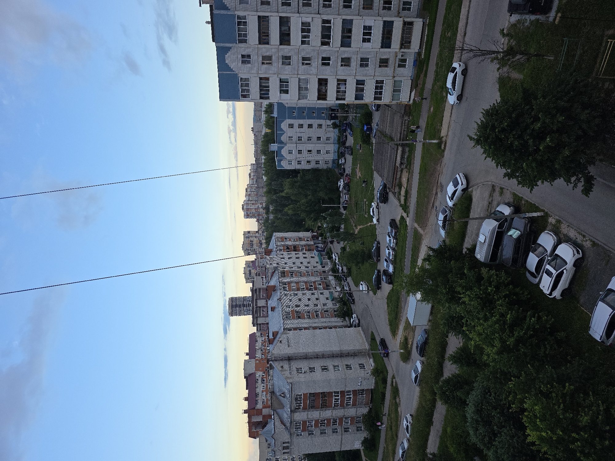 The image depicts a wide aerial view of a residential area in Novocheboksarsk, Russia, during what appears to be either dawn or dusk, given the soft lighting and clear sky with scattered clouds. Dominating the scene are several large, multi-story apartment buildings, characteristic of Soviet-era architecture, constructed with light-colored brick and concrete. Some buildings have blue accents on their upper floors.
The foreground and midground are filled with a dense canopy of green trees. Between the buildings and the trees, a network of paved roads and parking areas is visible. Numerous cars are parked in designated spots and along the streets, varying in color and type, including sedans, SUVs, and what appears to be a white van. A few individuals are seen walking along the sidewalks or roads.
In the background, more apartment buildings and some taller structures are visible, extending towards the horizon. The sky is a gradient of light blue with hints of yellow and orange near the horizon, suggesting the sun is low. Two thin, dark wires stretch diagonally across the upper portion of the image, likely utility lines. The overall impression is one of a typical urban residential neighborhood.