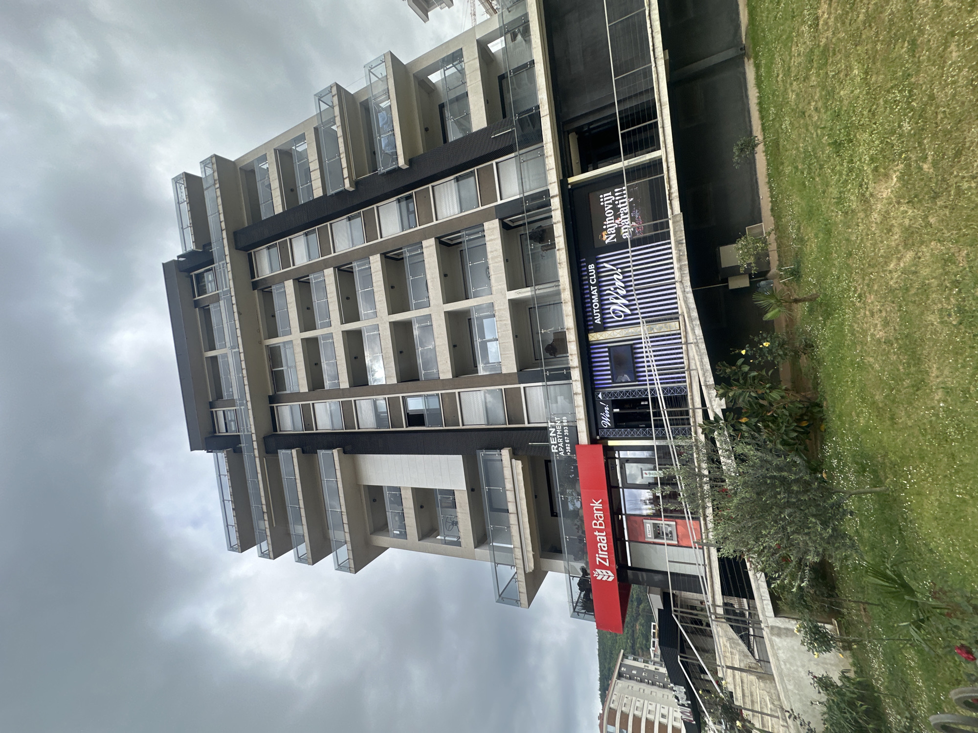 A modern, multi-story building stands prominently under an overcast, cloudy sky in this vertically oriented photograph. The architecture features a mix of beige concrete and dark grey or black panels, complemented by numerous balconies with clear glass balustrades.
The ground level of the building hosts commercial spaces. A bright red sign with the logo of "Ziraat Bank" is clearly visible. Above the bank, a white sign advertises "RENT APARTMENT" along with a Montenegrin phone number, "+382 67 393 989". Adjacent to the bank, an illuminated sign displays "AUTOMAT CLUB," "Win!", and "Najnoviji aparati!!!" (meaning "Newest machines!!!").
To the right of the building, a well-maintained green grassy area is present, dotted with small white flowers and various small trees and bushes. In the background, to the lower right, glimpses of another building and a verdant hill can be seen. The grey, cloudy sky suggests an overcast day. The scene depicts an urban or suburban environment in Budva, Montenegro, as indicated by the location cues. No individuals are visible in the image.