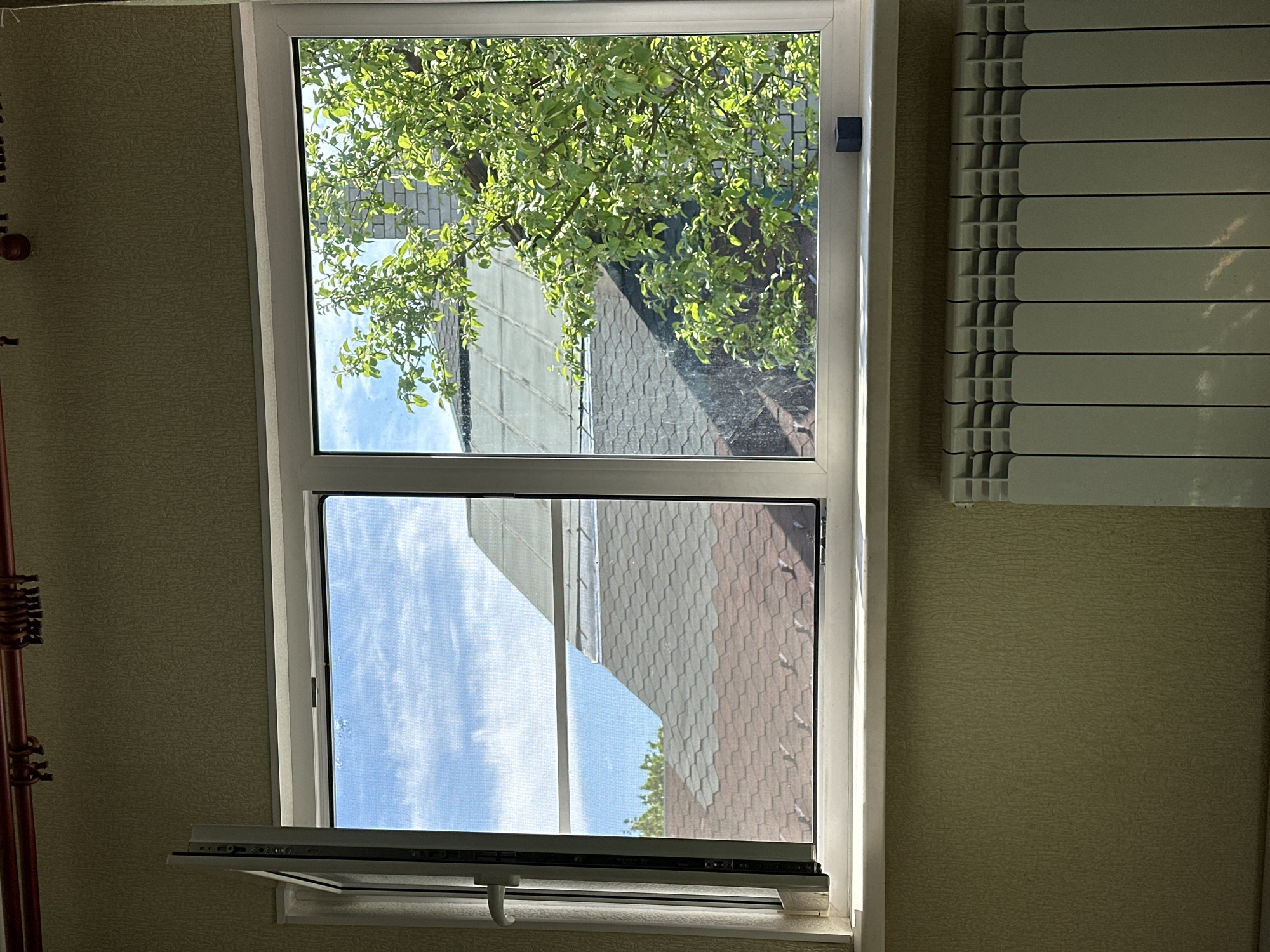 A white, partially opened window dominates the frame, offering a view to the outside. Through the upper pane, green foliage, a segment of a roof, and a blue sky can be seen. The lower pane presents a similar view of the sky and roof, obscured by a window screen. The open lower portion of the window features a handle and hardware. To the right of the window is a vertical white radiator. The surrounding wall is covered in patterned wallpaper. A blurry object is partially visible on the left side of the frame. The image was most likely taken in a residential or commercial building in Russia but without a specific address, it is impossible to identify the exact location in the city of Nizhny Novgorod. There are no people visible in the image.