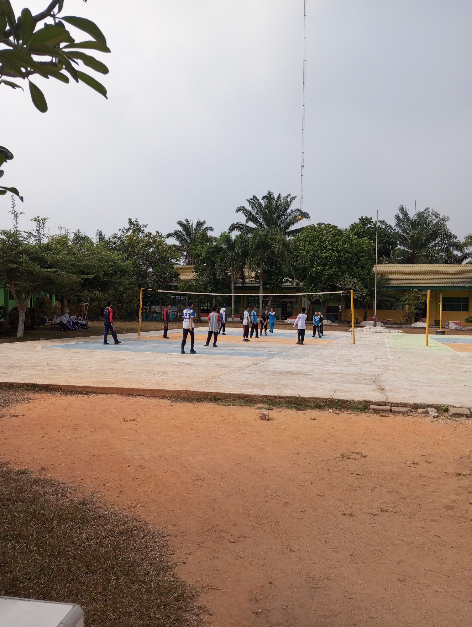 The image depicts an outdoor volleyball court in a schoolyard setting, likely in Pematang Reba, Indonesia, characterized by the presence of palm trees and tropical foliage. Several individuals, appearing to be students wearing athletic attire, are engaged in a game of volleyball. The court itself is paved and marked with colored lines indicating boundaries. In the background, a low-rise building with a yellowish-green facade and a distinctive tile roof is visible, suggesting it is part of the school infrastructure. Tall communication towers can be seen in the distance, against an overcast sky. The foreground shows a dirt and grass area, with a hint of a white object, possibly a bench or table, in the bottom left corner. The overall scene suggests a daytime activity taking place on school grounds.