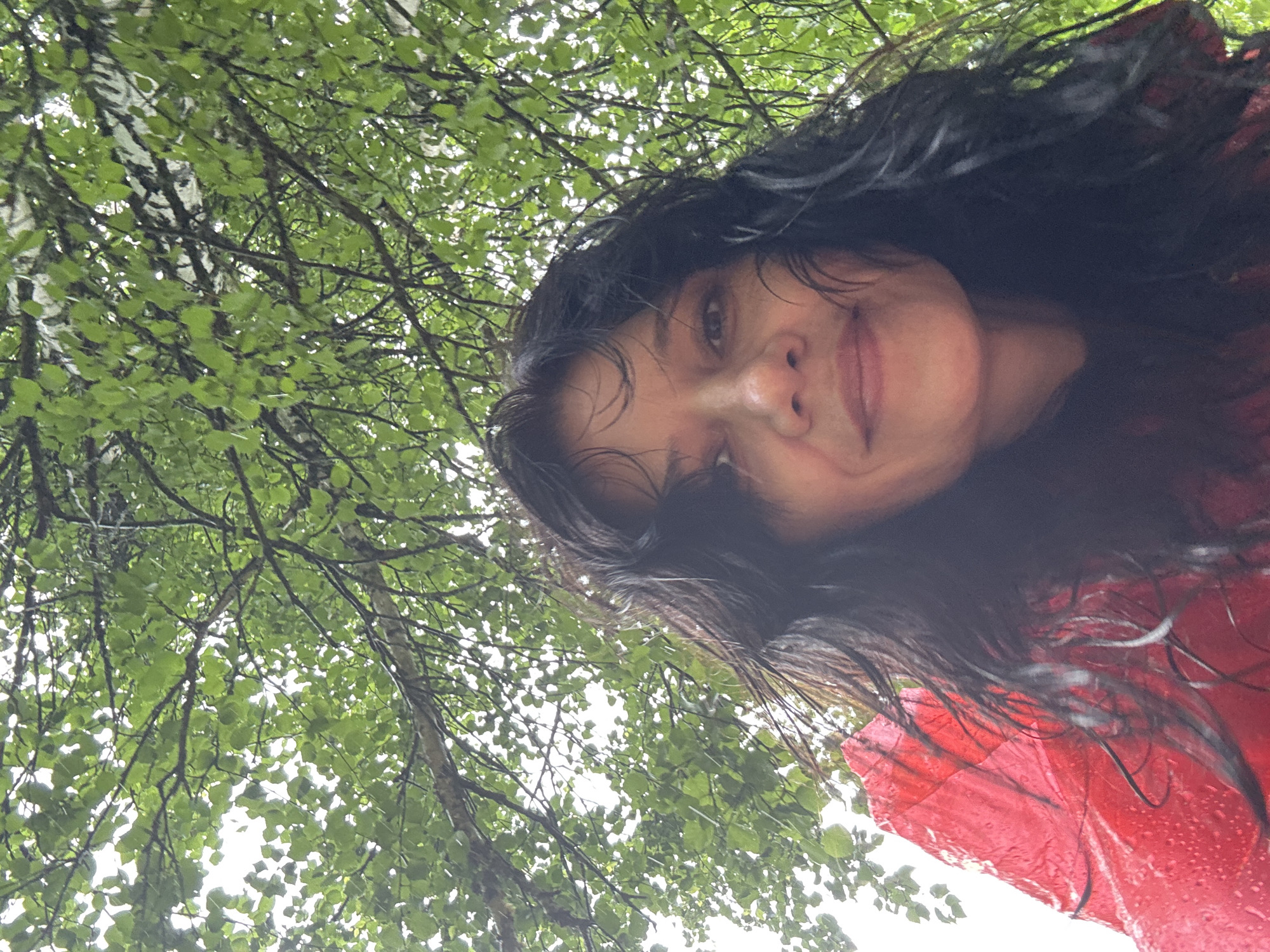 An outdoor, low-angle, close-up, selfie-style portrait of a person wearing a red hooded garment, possibly a raincoat, is shown. The person has long, dark, wet hair and is looking towards the camera with a slight smile. The background consists of a dense canopy of green leaves and branches from a tree, with dappled light filtering through. The overall impression is of a person under a tree in a slightly rainy or humid environment. The location is identified as Shumikhinskii, Russia.