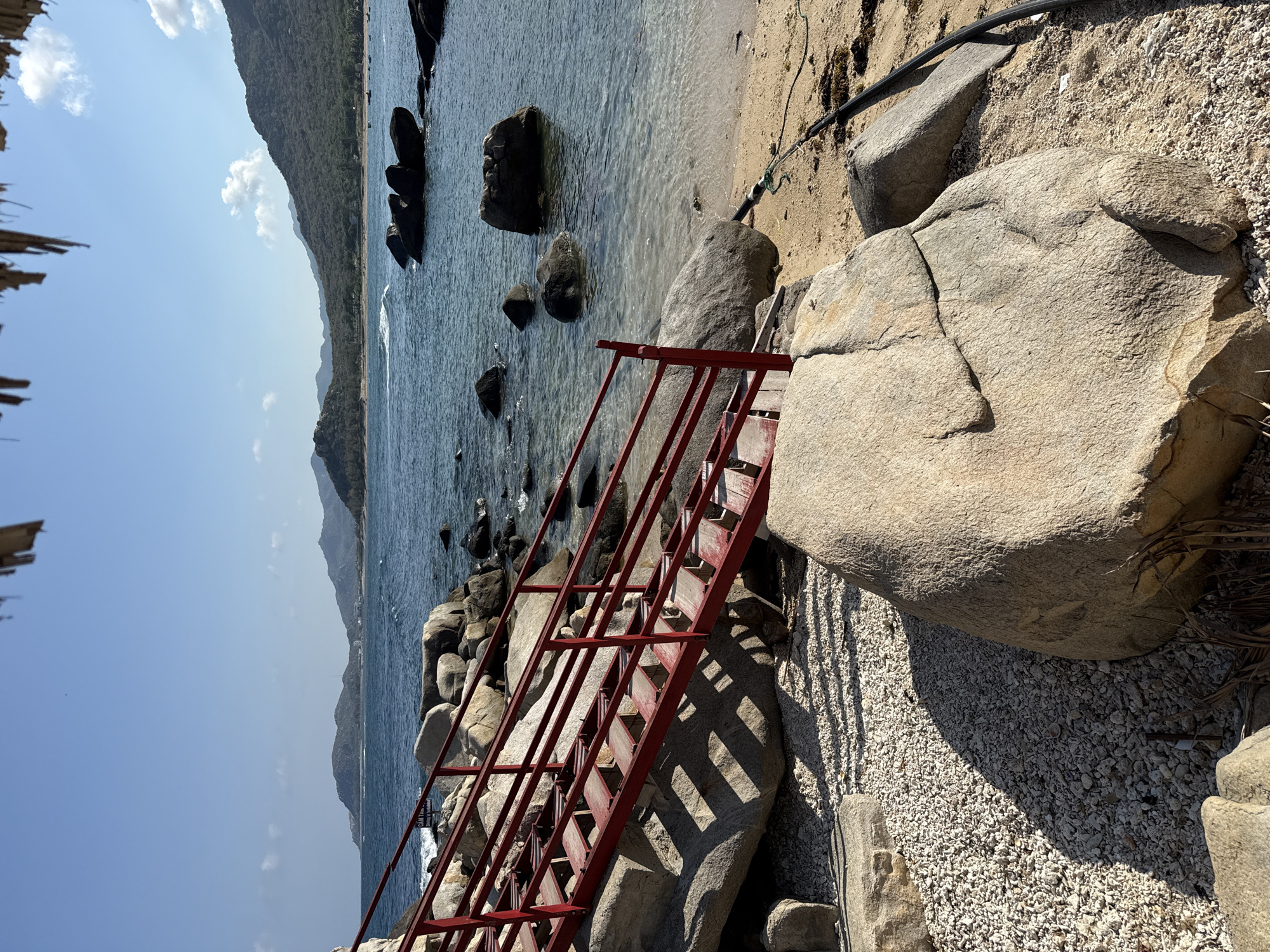 A coastal scene in Vinh Hai, Vietnam, features a rocky beach and coastline. Calm ocean water stretches to the base of a mountain range in the distance. A set of red metal stairs extends down from a rocky area toward the water. The stairs are partially obstructed by large boulders. The shoreline is a mix of sand and small shells. A black hose lies on the beach near the rocks.