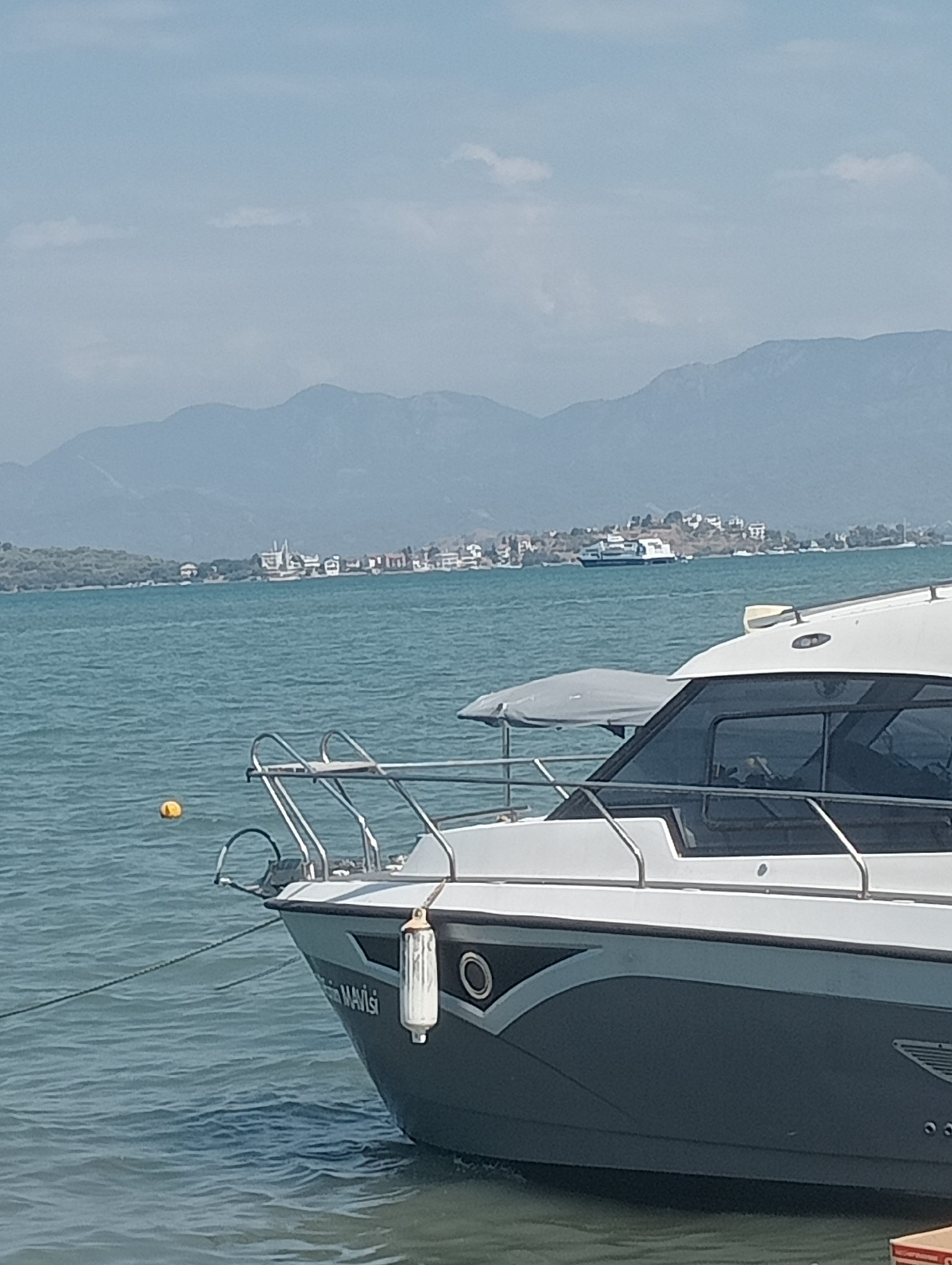 This is a medium-shot photograph of a boat moored in the turquoise waters of Fethiye, Turkey. The boat, predominantly white and grey, is in the foreground and takes up the right half of the frame. It has a sleek design with dark-tinted windows and chrome railings. The name "MAVİSİ" is visible on the side of the hull in white lettering. A white fender hangs from the side of the boat. In the distance, a low-lying coastline is visible with buildings and trees. The sea is calm with gentle ripples. Beyond the coastline, hazy blue mountains form the backdrop under a light blue sky with a few wispy clouds. In the mid-ground, a large white ferry or passenger boat is visible on the water, moving towards the right. The overall impression is one of a peaceful, sunny day in a coastal town.