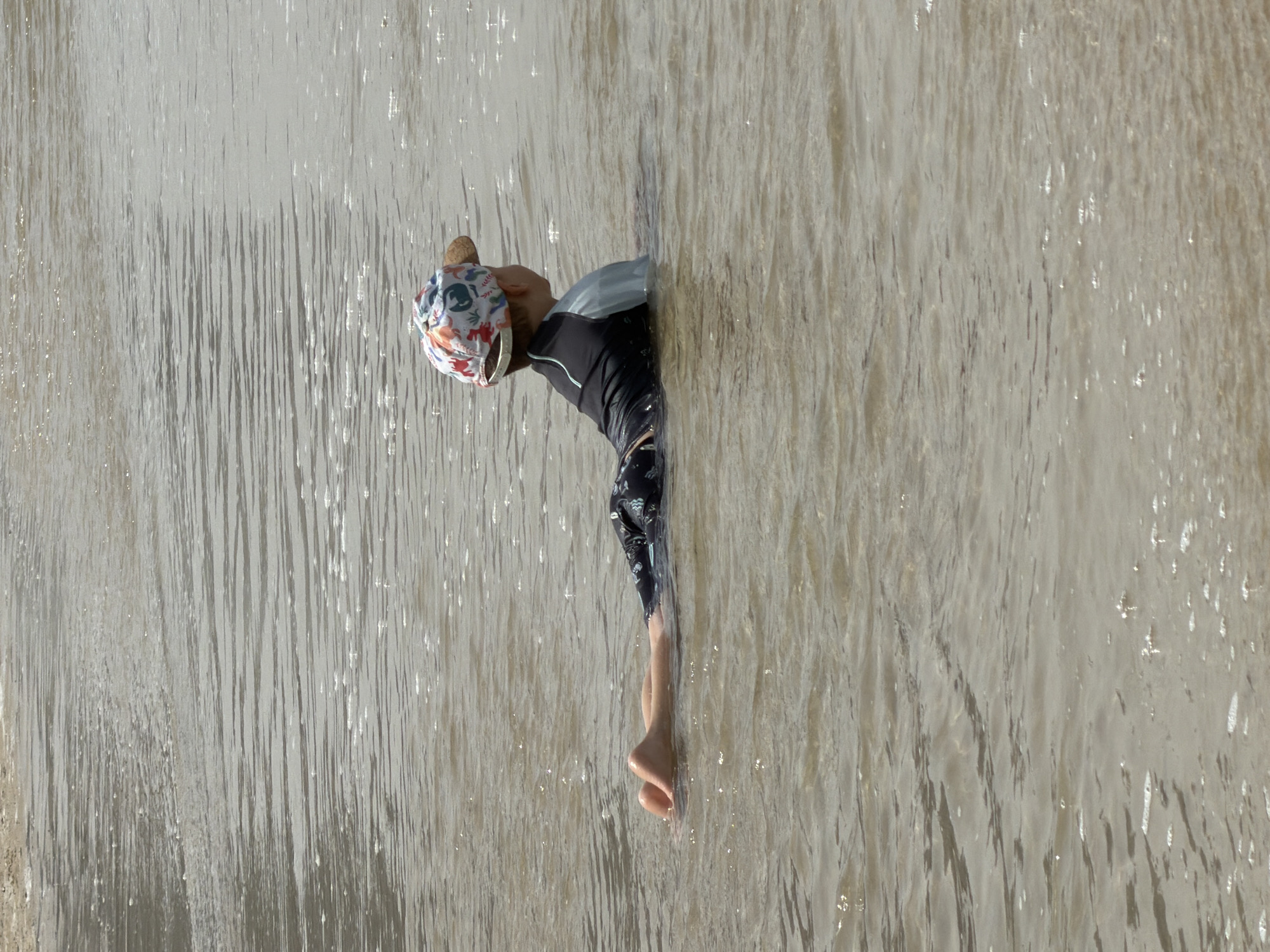 A person is standing in a body of water that appears to be cascading downwards, creating a curtain-like effect. The person is wearing a patterned cap, a black top, and patterned shorts. Their legs are submerged in the water, and only one foot is visible. The scene is presumably located in Tel-Aviv, Occupied Palestine. The water is a light brown color with streaks of white, giving the impression of movement and flow. The overall composition is somewhat abstract due to the angle and the obscuring effect of the water.