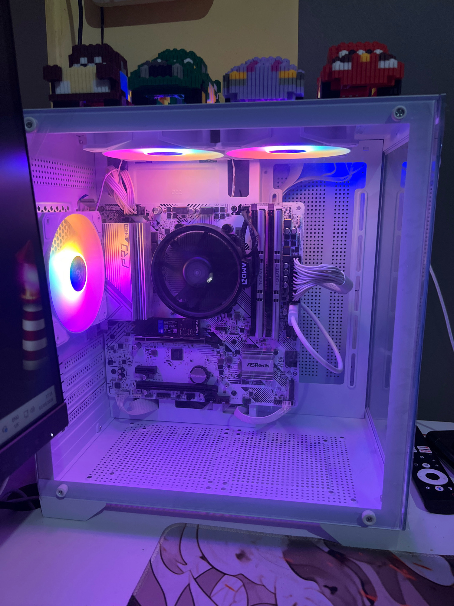 This is a close-up, eye-level shot of a white computer tower case with clear side panels, showcasing its internal components illuminated by purple and blue LED lighting. Inside, a motherboard with various components, including RAM sticks, a CPU cooler with a black fan, and cables, is visible. A large, circular RGB fan with rainbow lighting is mounted on the left side of the case. The computer is situated on a white desk, with a portion of a computer monitor visible on the left edge of the frame. In the background, on top of the computer case, are several small, pixelated figures made of building blocks, including a red car, a purple car, and a green dinosaur-like creature. The image does not provide any information that would allow for its location to be identified as Cimahi, Indonesia, nor does it depict any people, interactions, or actions. The scene is static and focused on the computer hardware.