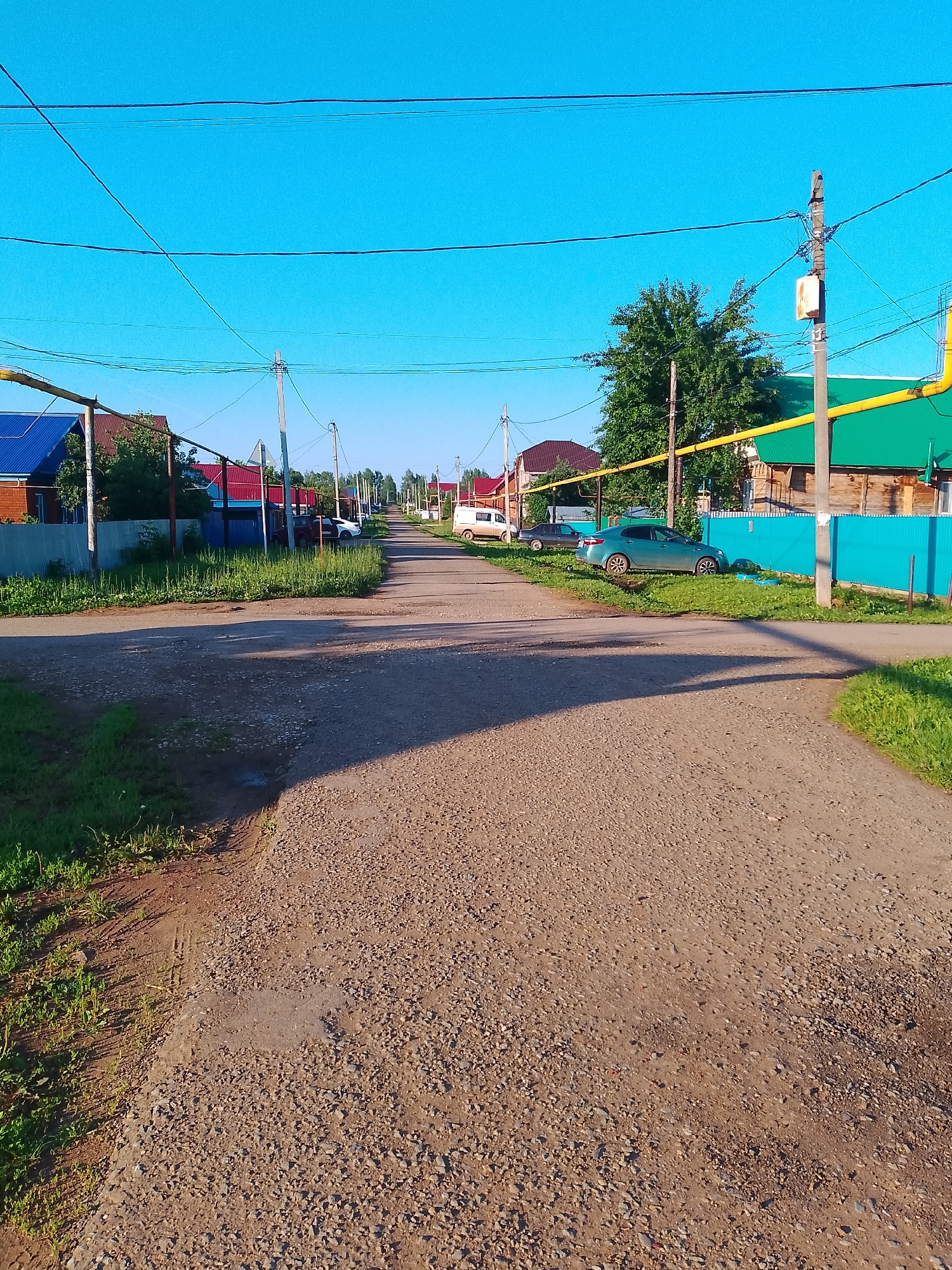 The image shows a view down a gravel road in a rural settlement, likely in the city of Krasnokholmsky, Russia. The road is lined with houses and fences on both sides, with trees and vegetation growing along the verges. Several cars are parked along the road, including a blue sedan, a white car, and a white van. Utility poles with electrical wires run along the sides of the road, and a yellow gas pipeline is visible overhead. The sky is clear and blue, and the sun is shining, casting shadows across the road. The scene appears to be a quiet, residential area.