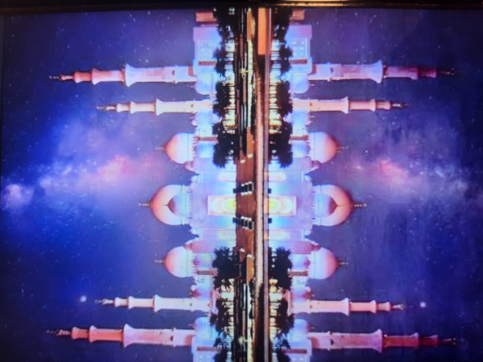 A digitally altered image depicts the Sheikh Zayed Grand Mosque, illuminated and reflected in water under a star-filled, nebula-like sky. The mosque, a white structure with domes and minarets, exhibits pink and orange hues, contrasting with the deep blue of the sky. The reflection shows a mirrored image of the mosque and the sky, with the water appearing dark. The scene is framed by what appears to be a television screen, with a black border visible on the right side. There are no people or animals visible in the image. The location is not Rosenheim, Germany, but rather Abu Dhabi, United Arab Emirates, the actual location of the Sheikh Zayed Grand Mosque.