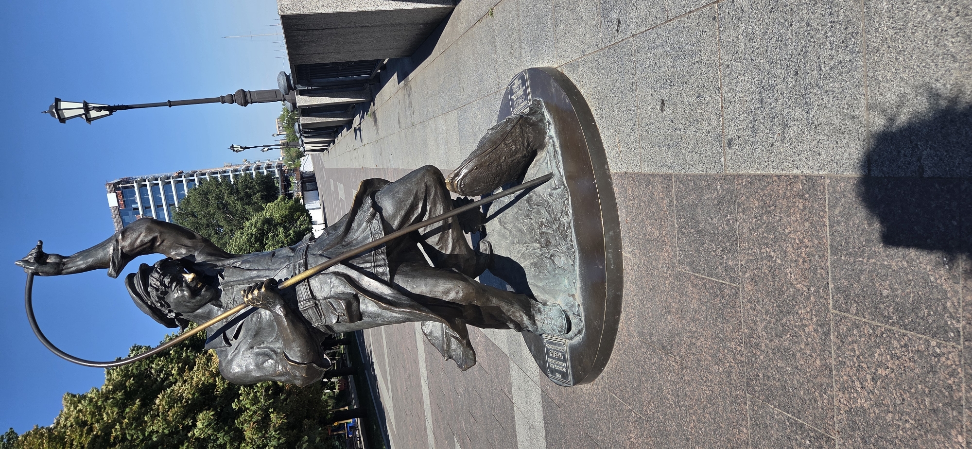 The image depicts a bronze sculpture of a man holding a fishing rod and a net, positioned on a circular stone pedestal. The sculpture is located in Rostov-on-Don, Russia, on the embankment of the Don River. The man in the sculpture is depicted in motion, with his left leg forward and his right leg bent, suggesting he is in the process of casting his net. He wears a cap, a long coat, and trousers, and his face is rendered with a determined expression. A large fish is caught in the net he holds.
The sculpture is situated on a paved walkway, with granite paving stones in varying shades of gray and brown. In the background, a modern high-rise building with reflective windows is visible, under construction with scaffolding. Several lampposts line the walkway, and a black railing runs along the embankment, separating the walkway from the river. Trees with lush green foliage are present on the left side of the image. The sky is clear and blue, indicating a sunny day. The shadow of the sculpture and potentially a nearby object is cast on the paved ground. The overall scene portrays an outdoor public space with a focus on a prominent piece of art, set against an urban backdrop with elements of nature.