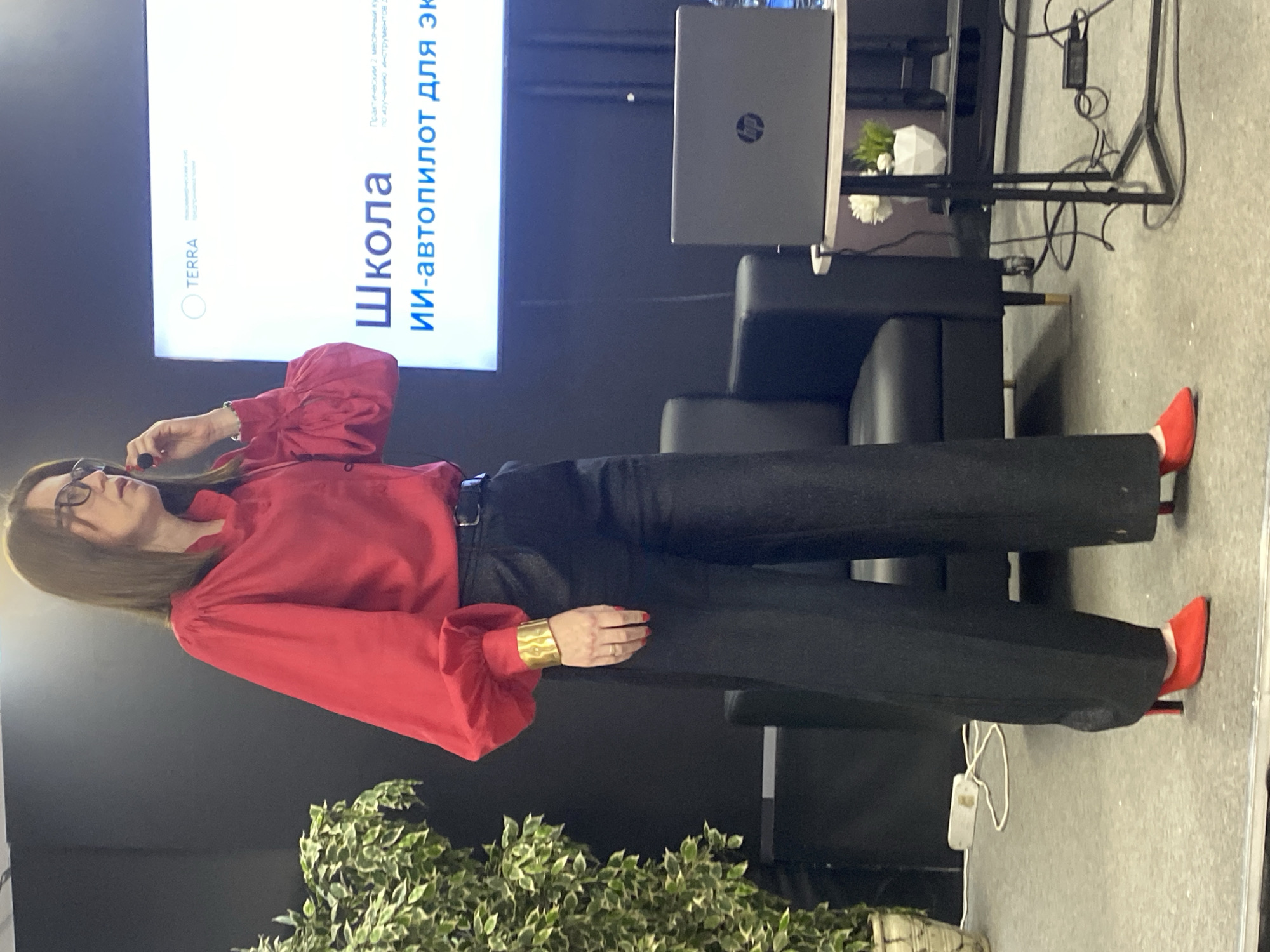 A woman wearing a red blouse, black pants, and red shoes is standing in front of a screen that says "Школа ИИ-автопилот для эк" (School AI autopilot for ex). She is wearing glasses and has a gold bracelet on her wrist. There is a laptop and a chair behind her. There are plants in the foreground. The location is Unknown, Unknown.