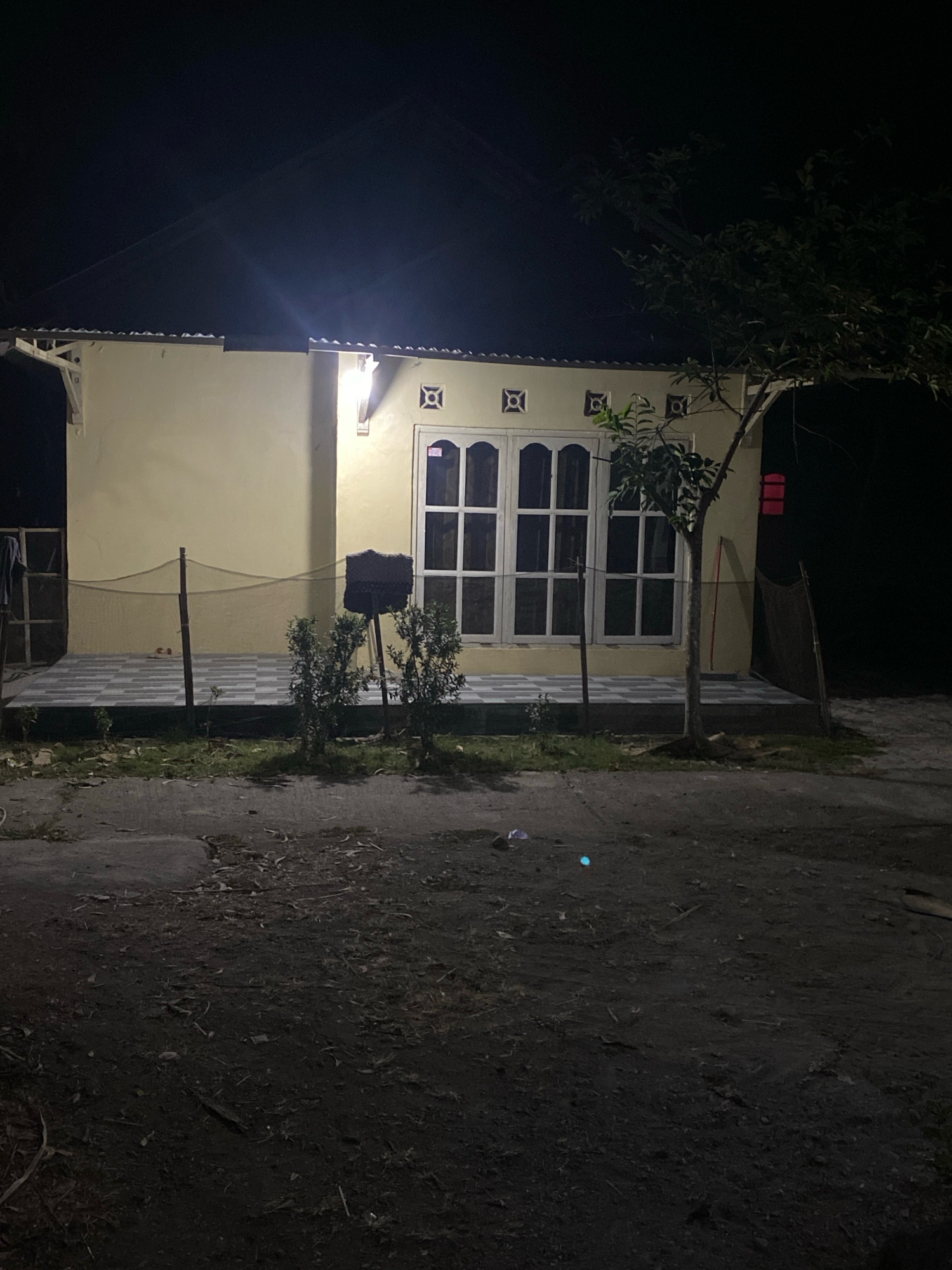 The image shows a single-story building with a yellow facade and a tiled front porch at night in Planjan, Indonesia. A large, multi-paneled window is visible on the front of the building. A young tree stands to the right of the window, and several small bushes are planted on the porch. The area in front of the building is a dirt road covered in debris. A fence surrounds the porch. The scene is lit by artificial light, casting shadows.