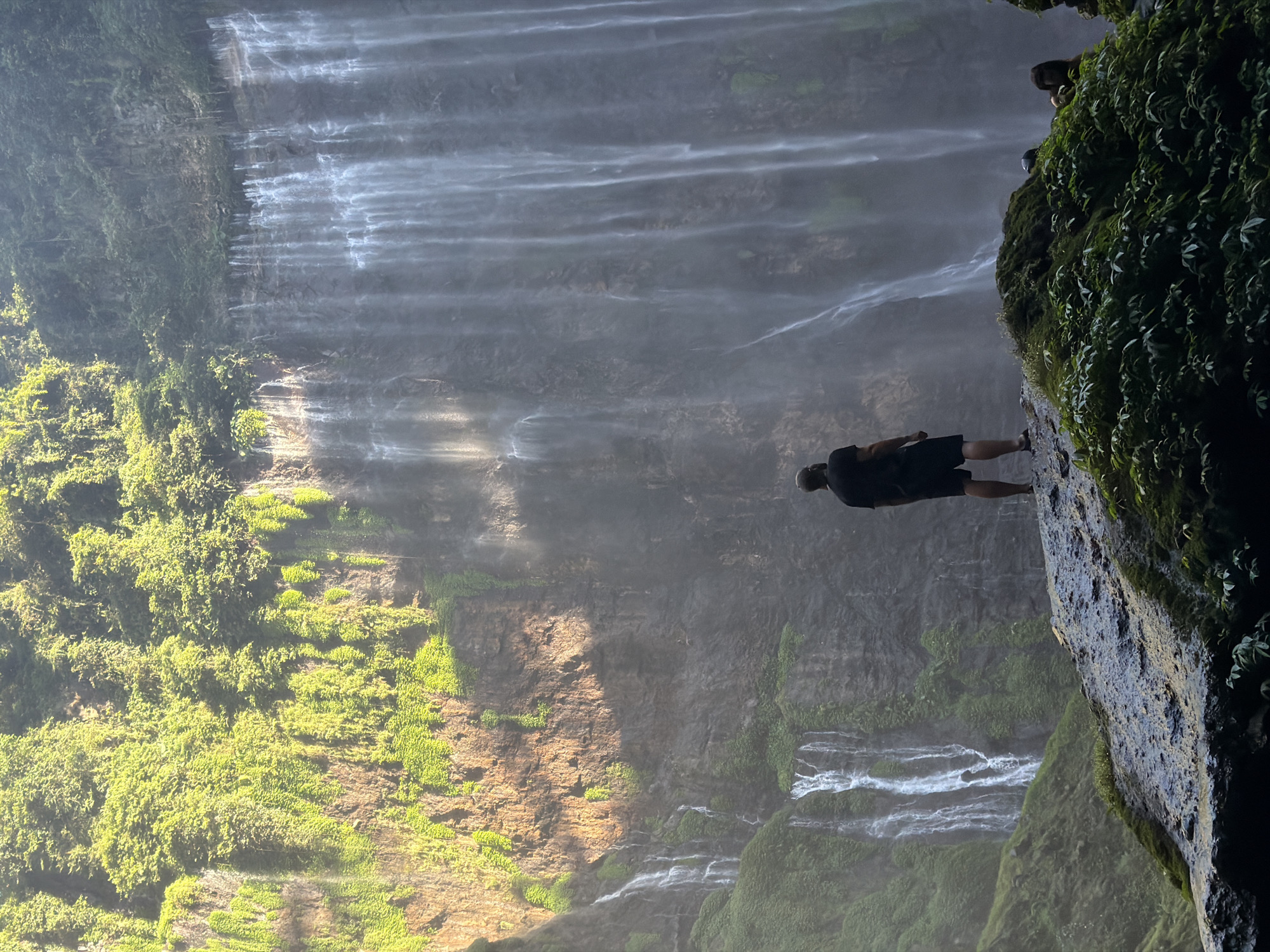 The image depicts a majestic waterfall cascading down a steep, rocky cliff face covered in lush green vegetation. The scene is set in Sidomulyo, Indonesia. In the foreground, a lone individual stands on a moss-covered rock, facing the waterfall, with their back to the camera. The person is wearing a black t-shirt and black shorts. The waterfall is the dominant feature, with multiple streams of water flowing down the cliff. The sunlight illuminates parts of the cliff face and the mist rising from the waterfall. There are other individuals visible in the lower right corner of the image, partially obscured by foliage, appearing to be observing the waterfall. The environment is natural and verdant, characterized by the presence of water, rocks, and dense plant life.