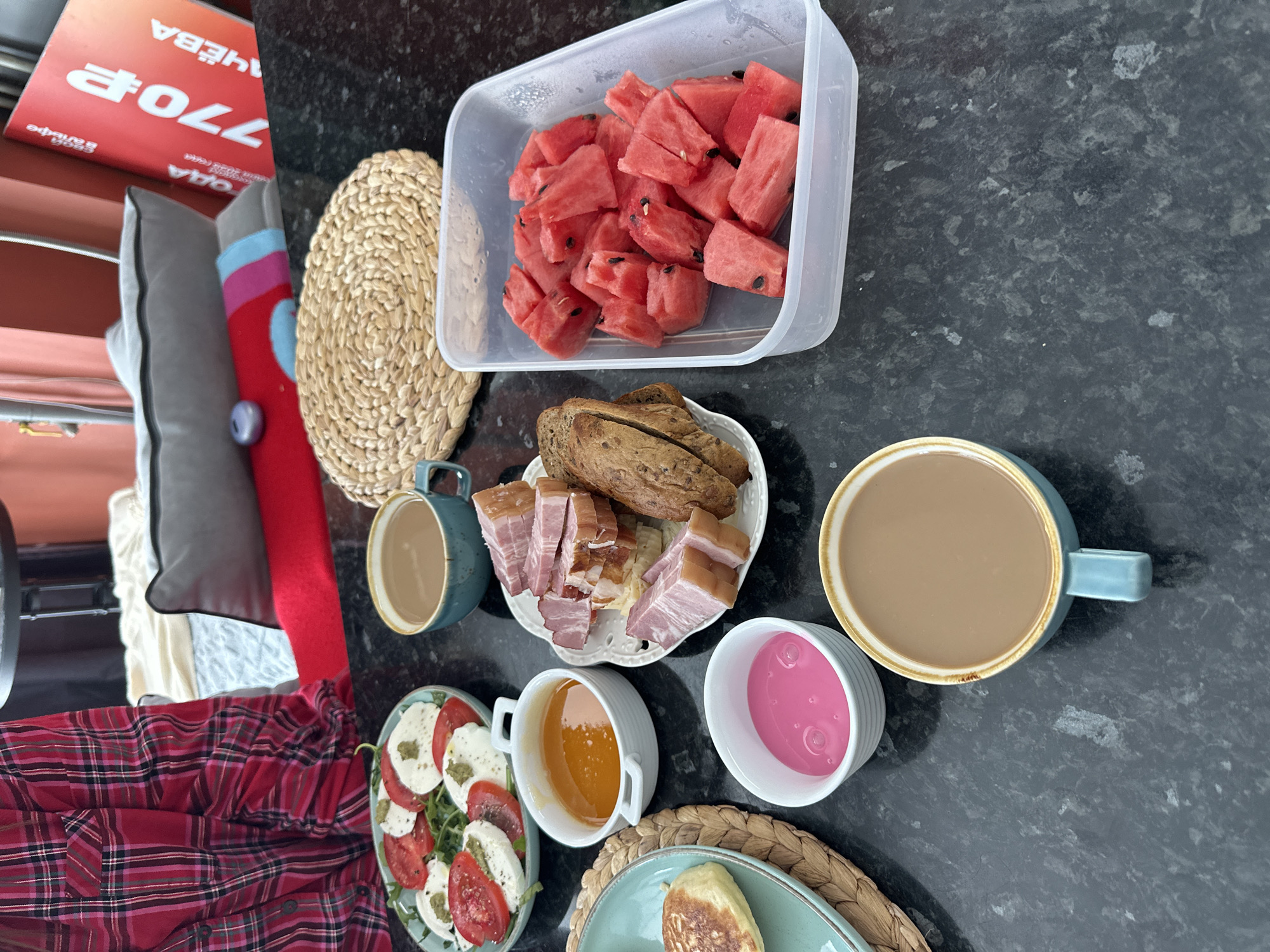 The image displays a breakfast spread on a dark granite countertop, likely in a home environment in Sochi, Russia. The food items include a salad of sliced tomatoes and mozzarella cheese, a small dish of orange jam or sauce, sliced smoked meat with some bread, a container of cut watermelon, and two cups of coffee. There is also a pancake or fritter on a teal plate and a small bowl of pink yogurt or dessert. In the background, a person wearing a red and black plaid shirt is partially visible. A red and white promotional sign with Russian text is also present. The overall scene depicts a casual breakfast setting.
