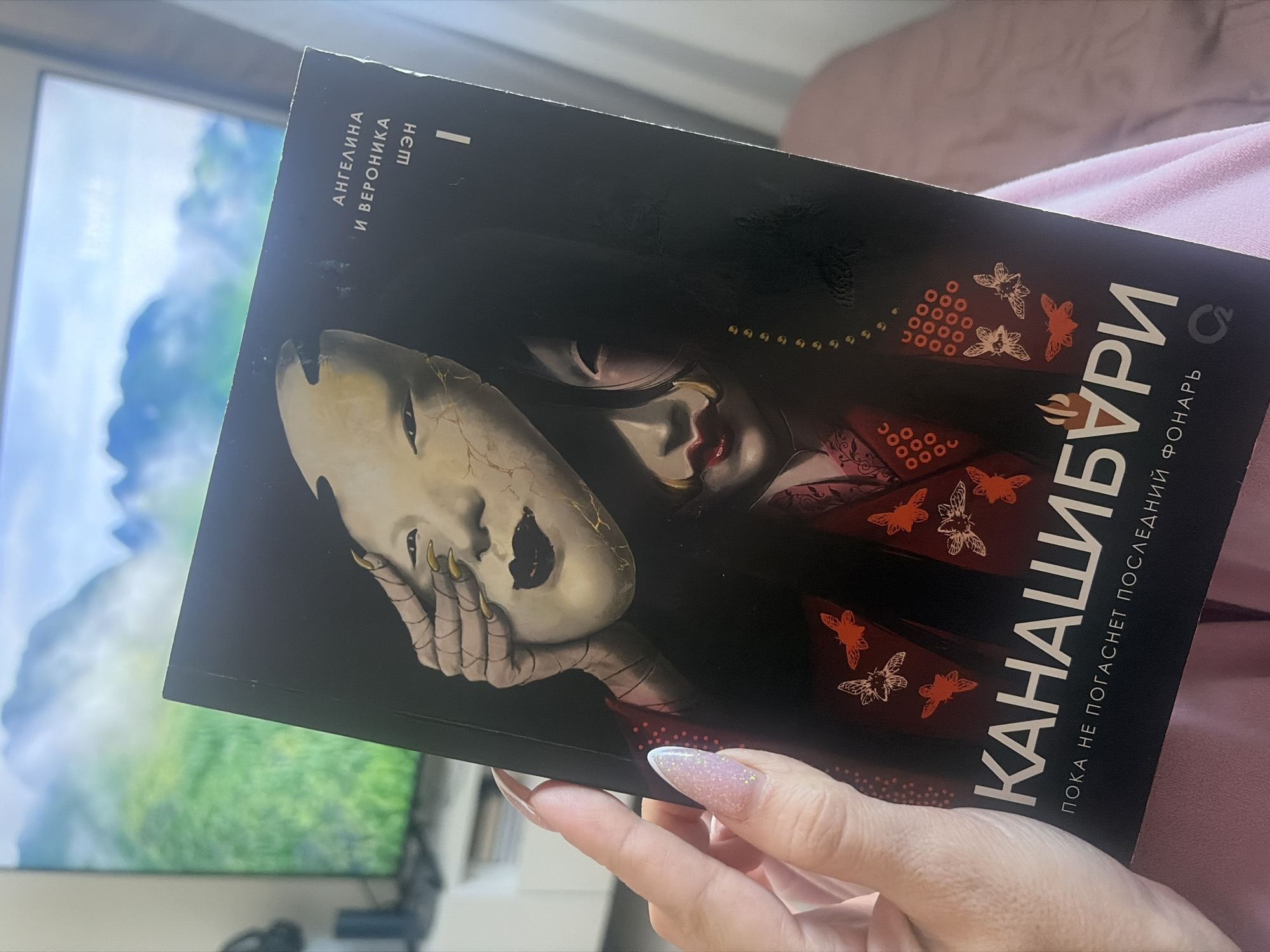 A right hand with long, almond-shaped fingernails, painted with sparkling light pink and white glitter polish, holds a book open. The book's cover is dark, featuring the Cyrillic title "КАНАШИБУРИ" (KANASHIBURI) in large white letters. Beneath the title, a tagline reads "ПОКА НЕ ПОГАСНЕТ ПОСЛЕДНИЙ ФОНАРЬ" (UNTIL THE LAST LANTERN GOES OUT). The authors, "АНГЕЛИНА И ВЕРОНИКА ШЭН" (ANGELINA AND VERONIKA SHEN), are listed vertically along the top left edge.
The cover art depicts a stylized figure, likely a woman with long black hair, holding a cracked, pale, mask-like object with golden accents over her face. Her visible lower face shows pale skin and red lips, while the hand holding the mask has long, gold-painted nails. The dark background of the cover features scattered patterns of red circles and various white and orange butterflies or moths. The figure appears to be dressed in a red, possibly kimono-style garment.
The image is captured indoors during daytime, with natural light illuminating the scene. In the out-of-focus background, a large screen, possibly a television or monitor, displays a soft blue and green image, suggesting a landscape or digital art. A soft pink fabric, possibly a blanket or clothing, is visible behind the hand. The book's cover artwork suggests an East Asian, specifically Japanese, theme. The scene provides a relaxed indoor setting, potentially in Saint Petersburg, Russia.
