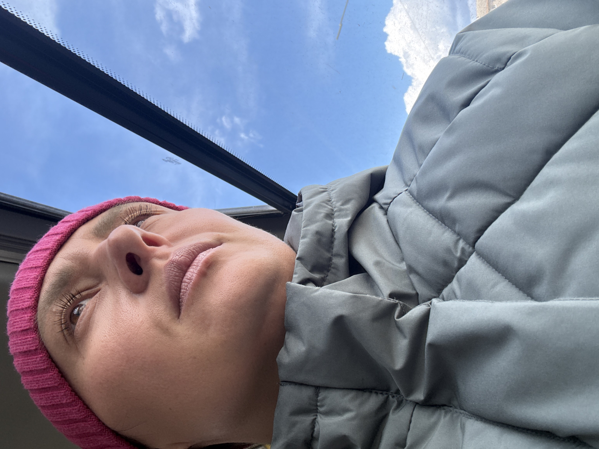 The image, captured from a low angle and rotated counter-clockwise, presents a close-up of a person looking upwards through a vehicle's sunroof or large window. The person occupies the lower-left portion of the frame (when oriented upright, they would be on the right, looking up and slightly left), while the upper-right shows a bright blue sky with scattered white clouds.
The subject is wearing a bright pink knitted beanie and a light grey quilted or puffer jacket. Their face is visible in profile, showing their nose, lips, and one eye gazing upwards. The expression appears neutral, perhaps serene or contemplative, as they look towards the sky. Details like long eyelashes are discernible.
Above the subject, a dark, rigid frame, likely part of the vehicle's interior structure or a window seal, runs diagonally across the transparent panel. This frame features a dotted pattern, possibly a frit band found on car windows.
The bright lighting and clear sky indicate it is daytime with fair weather. The setting is inside a vehicle, likely a car, with the person looking out through a roof window. No specific activity is depicted beyond observing the sky. There is no visible text in the image. The location is Chaikovsky, Russia.
